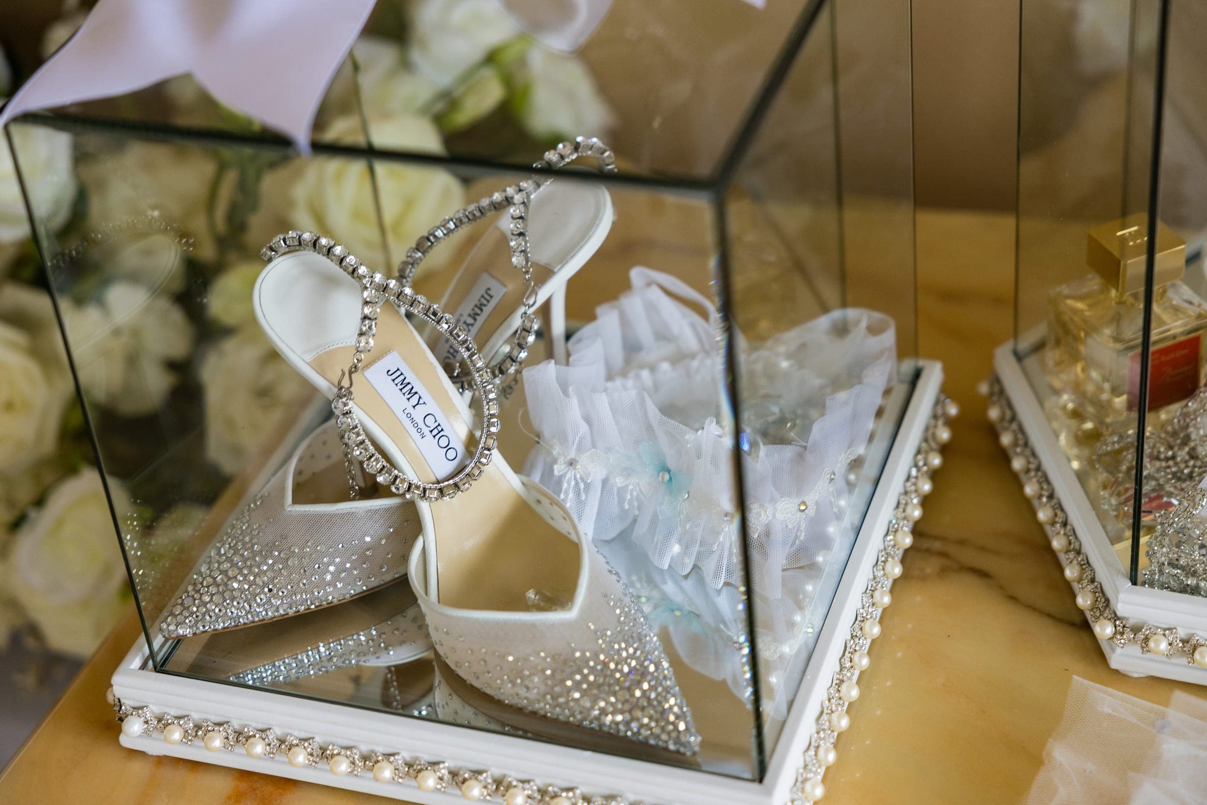 Crystal-embellished Jimmy Choo heels and a lace garter sit inside a mirrored display case beside white roses.