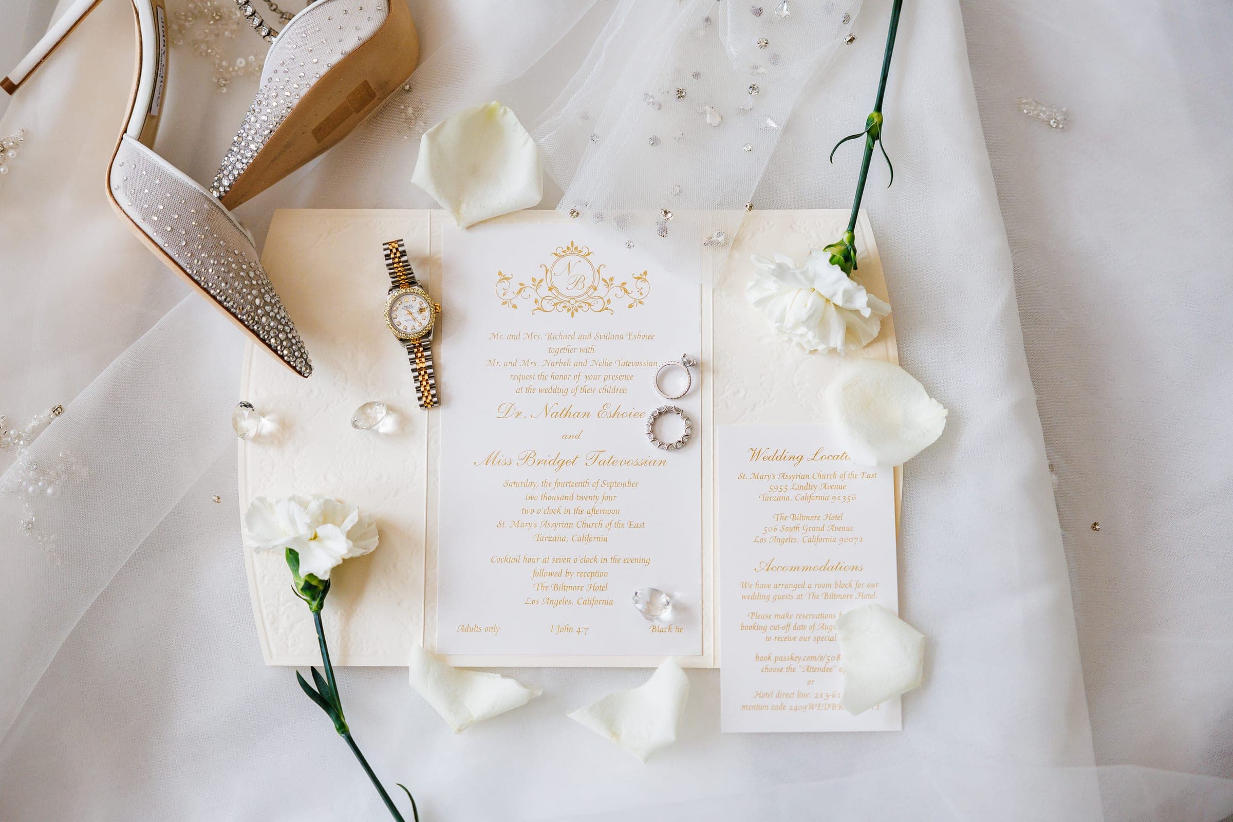 Wedding invitation, sparkling heels, gold watch, rings, and white flowers arranged on a soft veil backdrop.