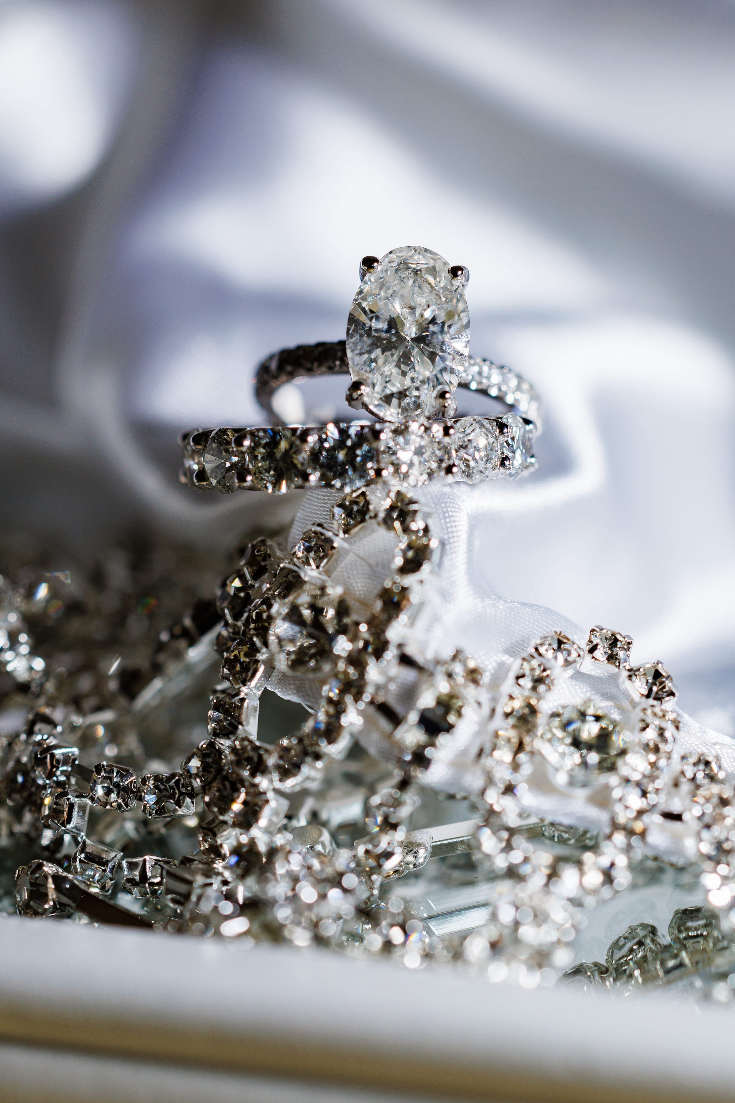 A diamond solitaire engagement ring rests atop a wedding band above sparkling rhinestone jewelry on white satin.