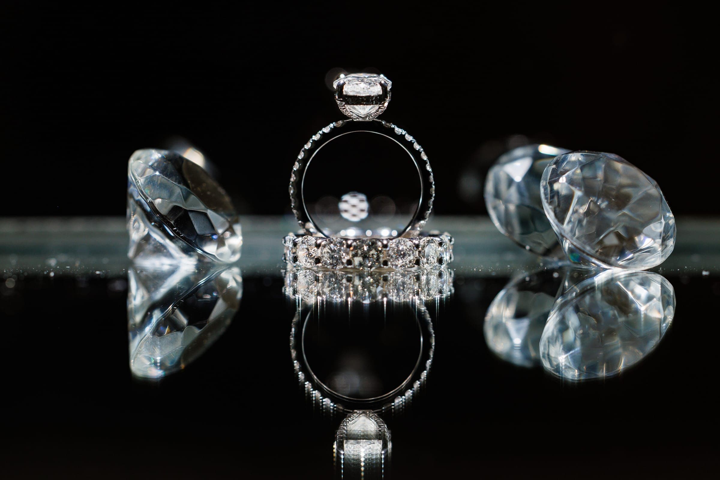 An engagement ring and diamond band reflected on black glass beside two large crystal gems.