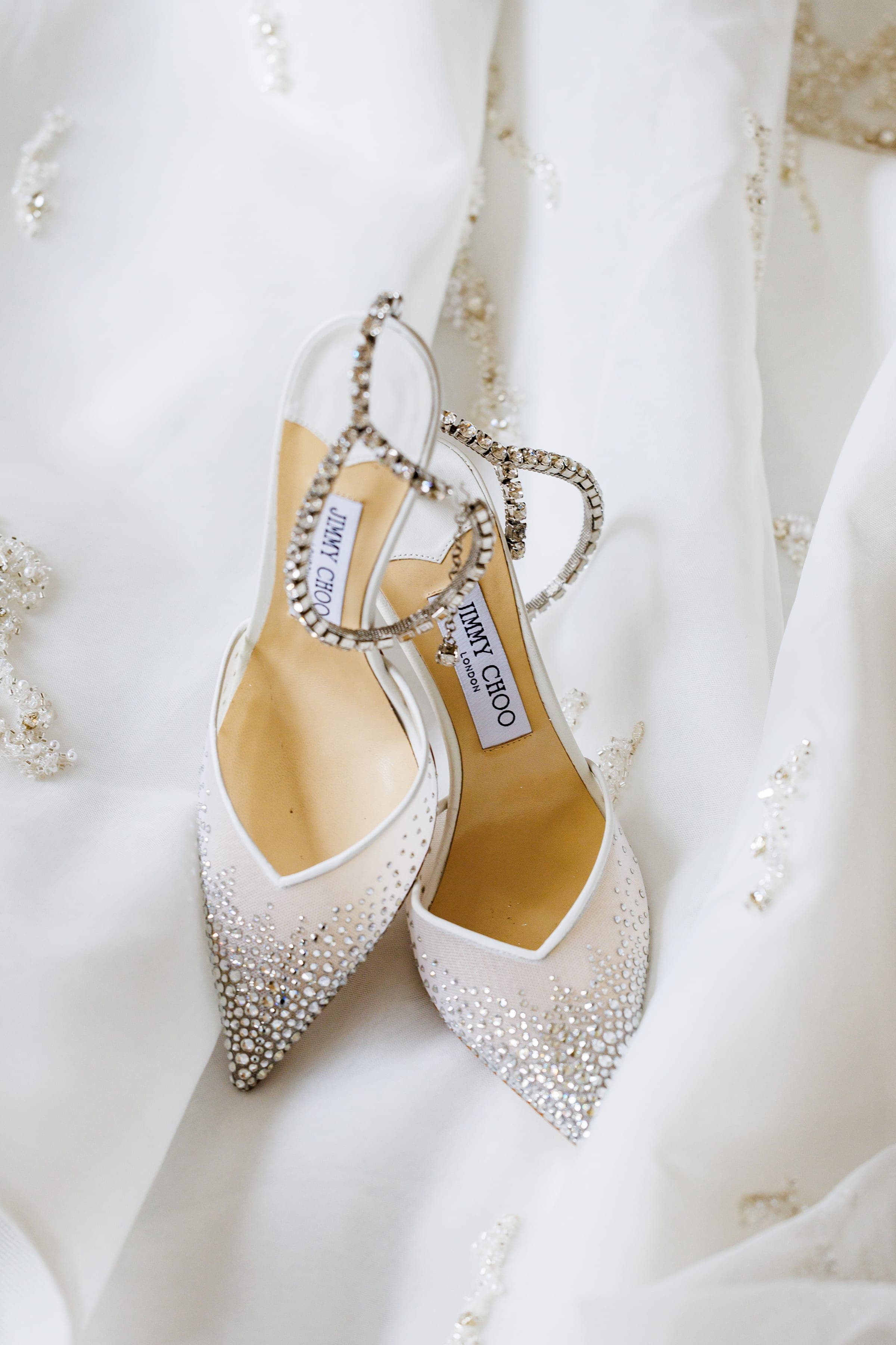 White Jimmy Choo crystal-embellished heels with rhinestone straps lie on ivory beaded fabric.