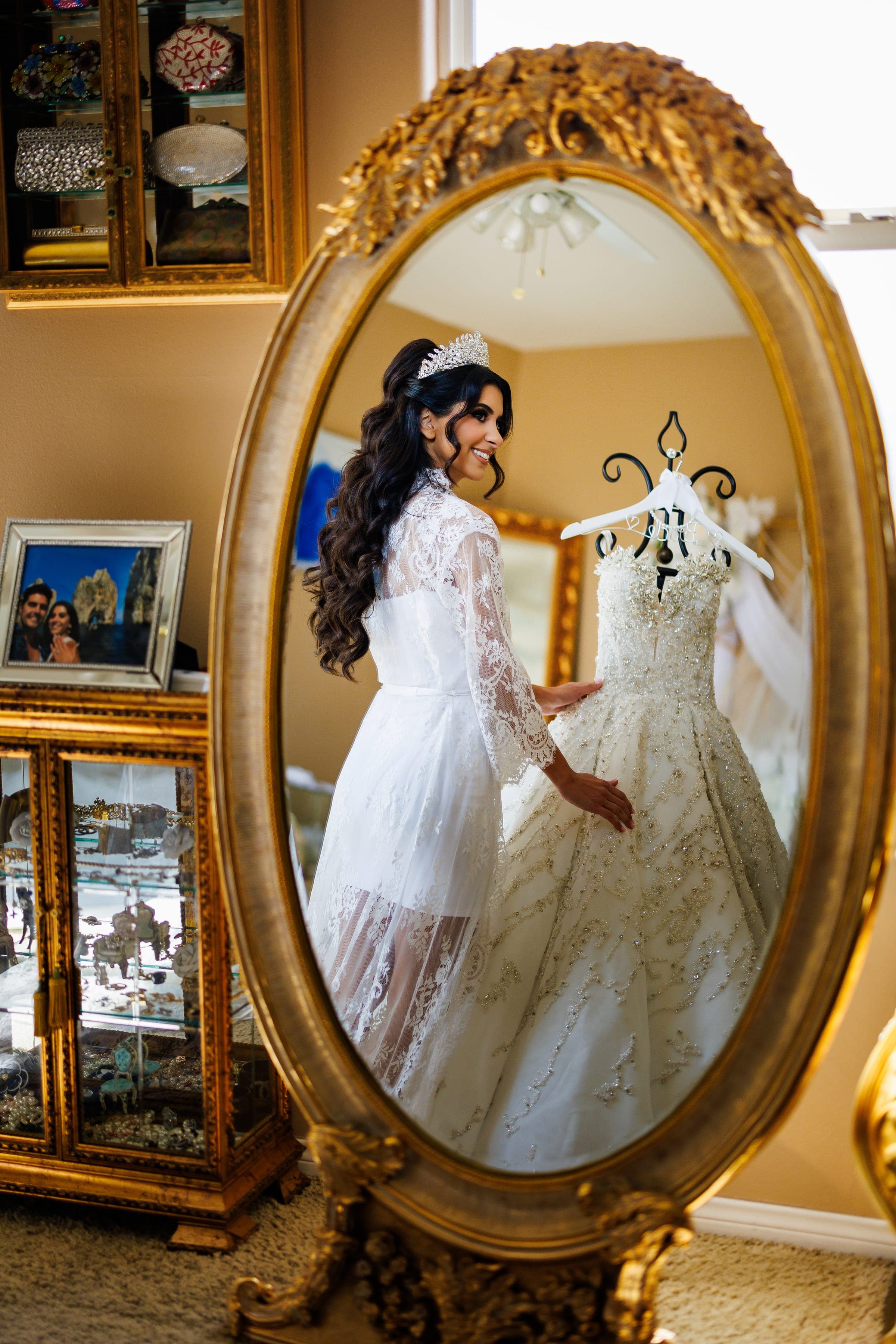 Bride in a lace robe and crown smiles beside her beaded gown, reflected in an ornate gold mirror.