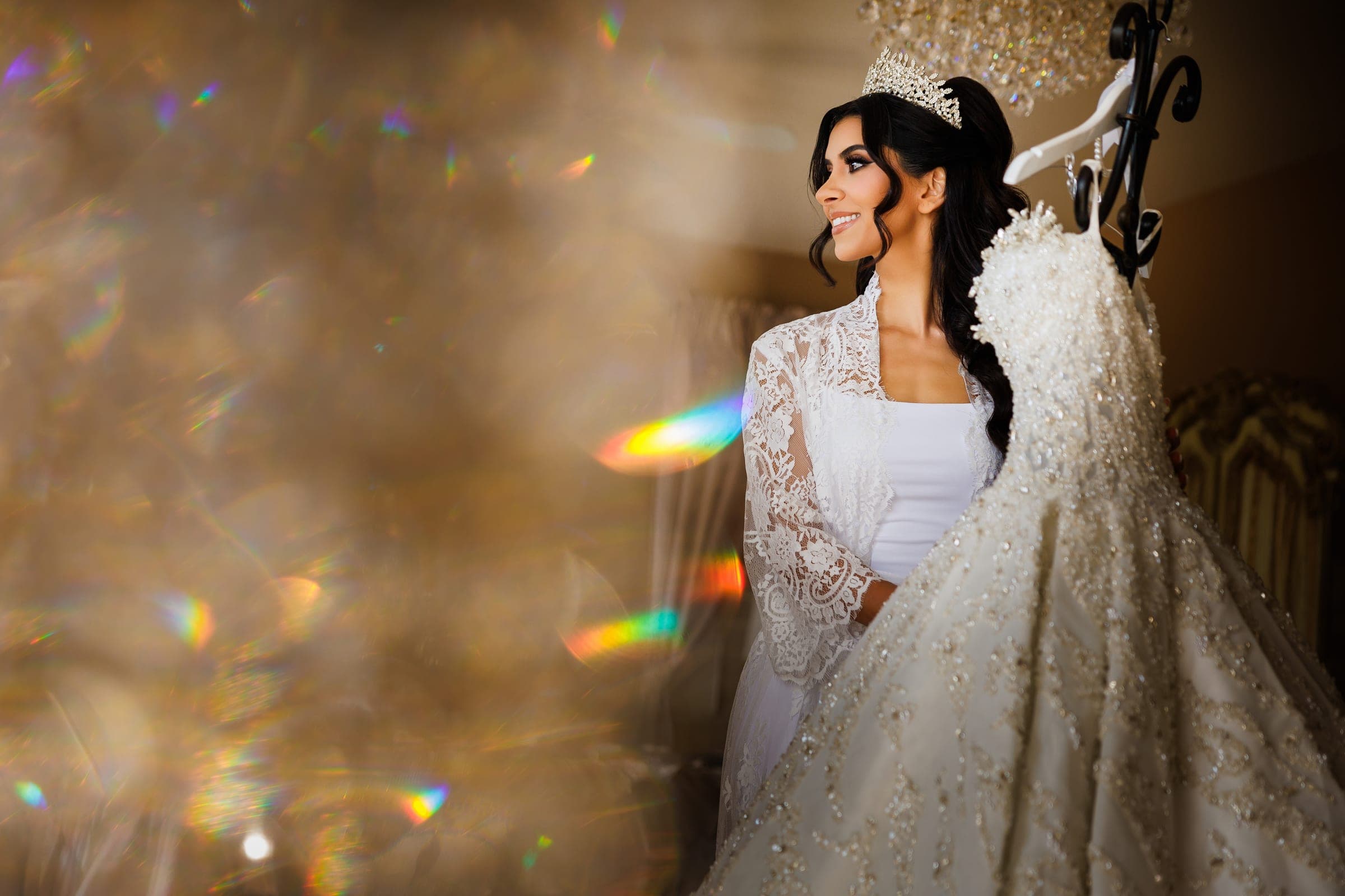 Bride in lace robe and crown smiles at her sparkling gown amid warm golden light and rainbow flares.
