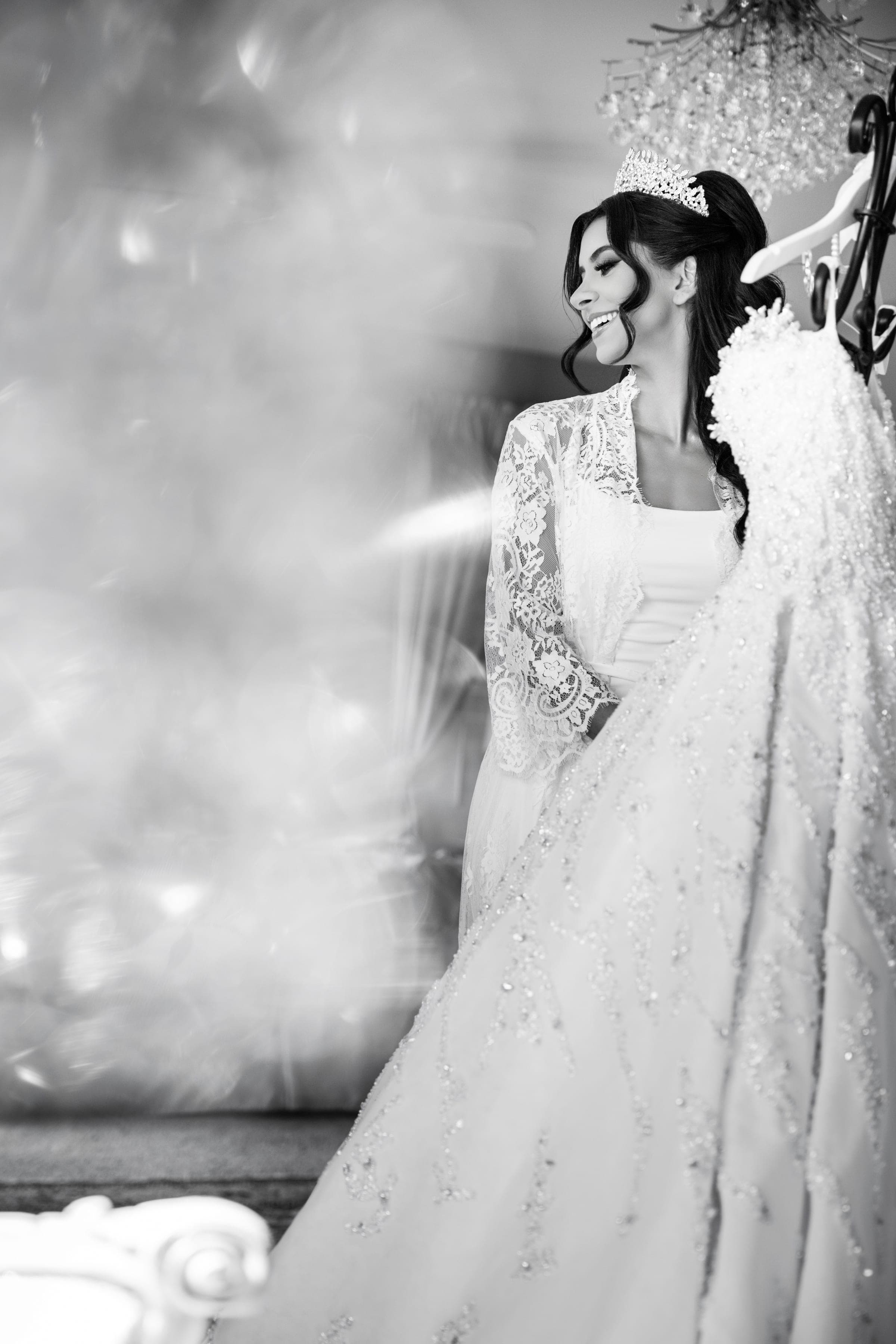 Black and white portrait of the bride in lace robe and crown smiling beside her beaded gown.