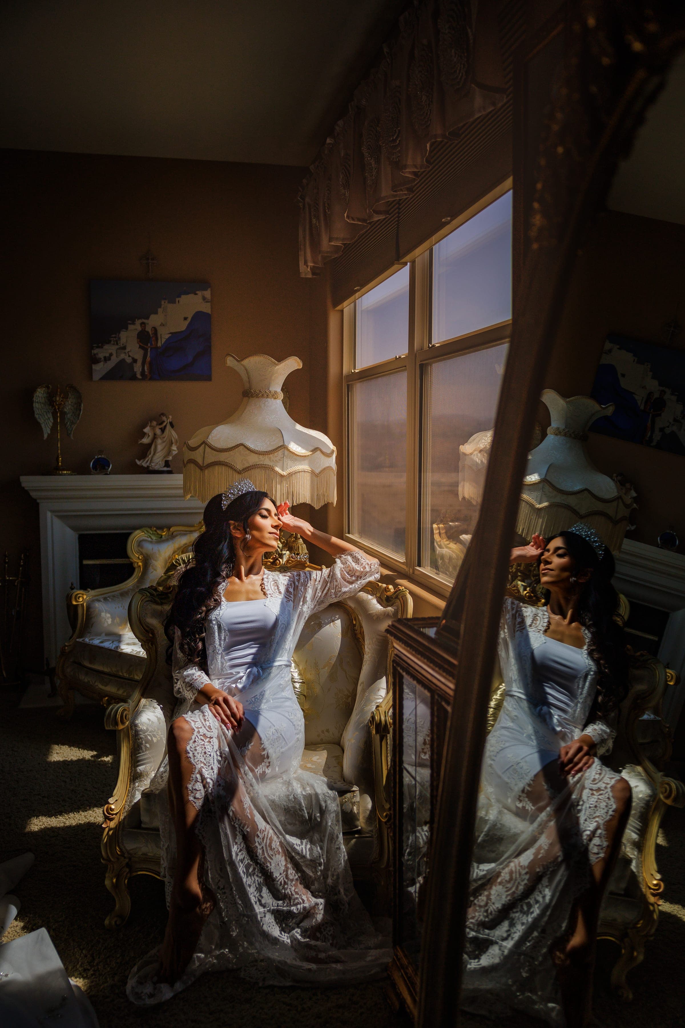 Bride in white lace gown and crown sits on gilded chair by a sunlit window, reflected in a mirror.