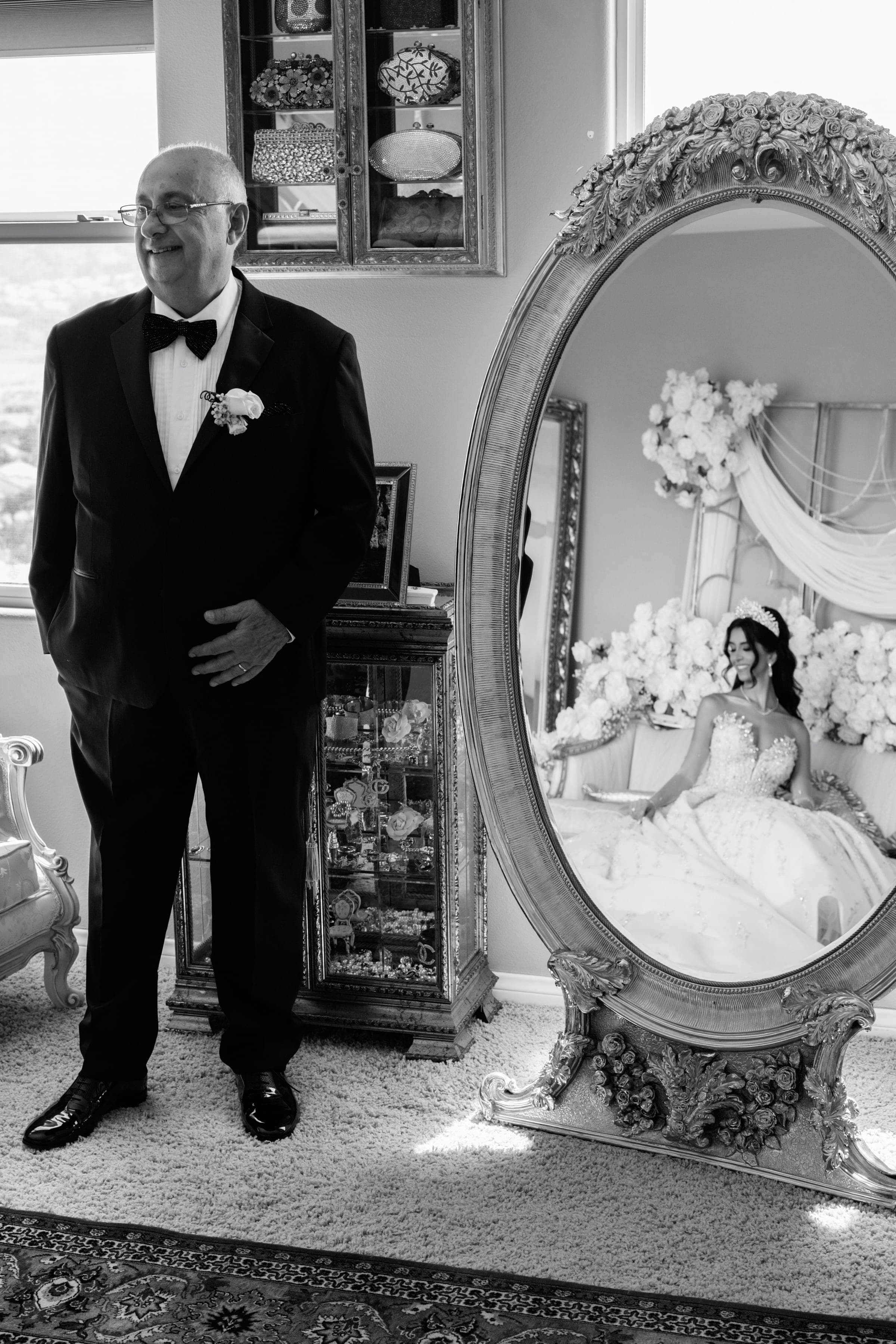 Smiling father in tuxedo stands beside an ornate oval mirror reflecting the seated bride in her gown.