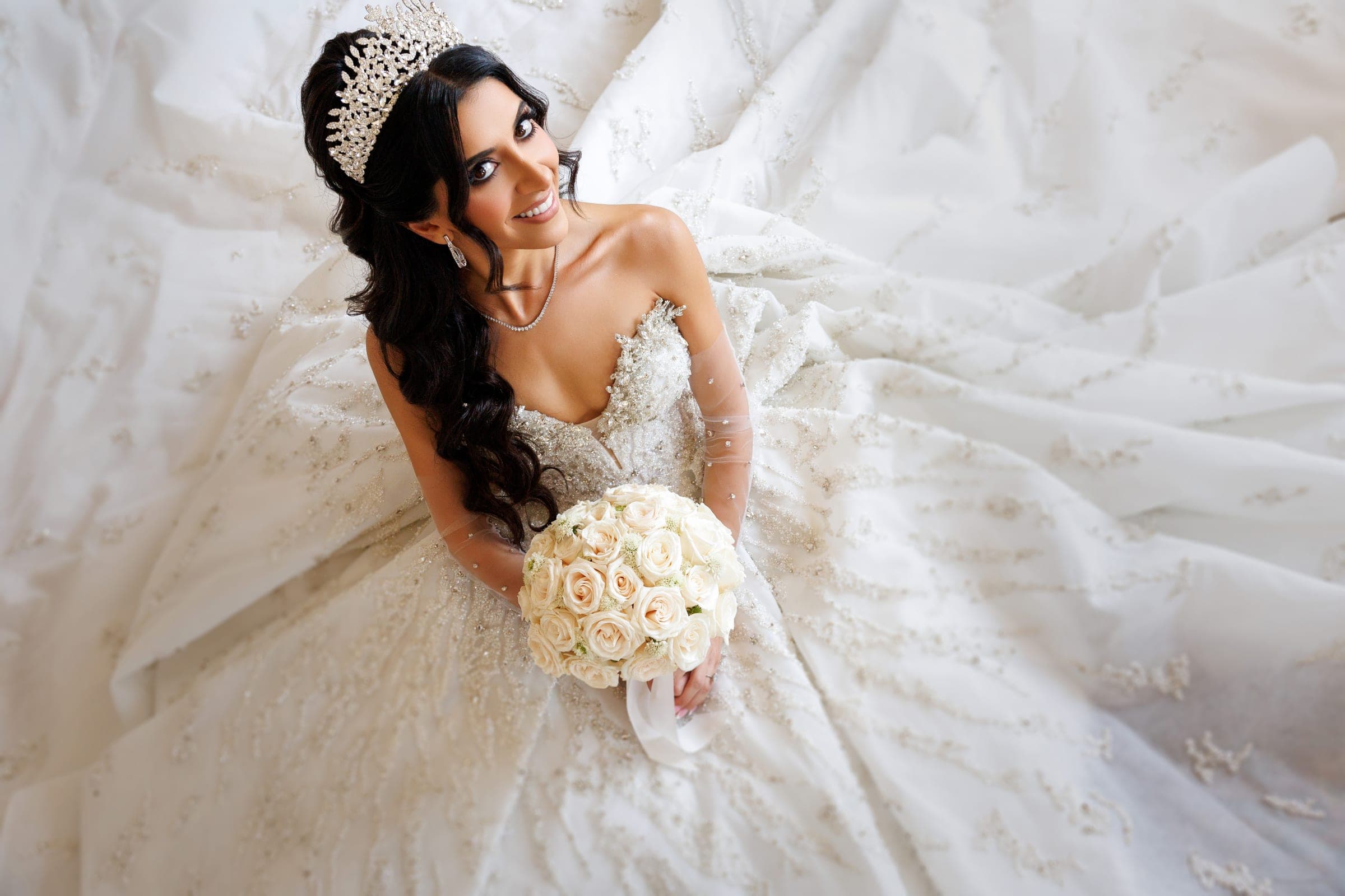 Bride in beaded gown and crown holds a cream rose bouquet, smiling up amid flowing white fabric.