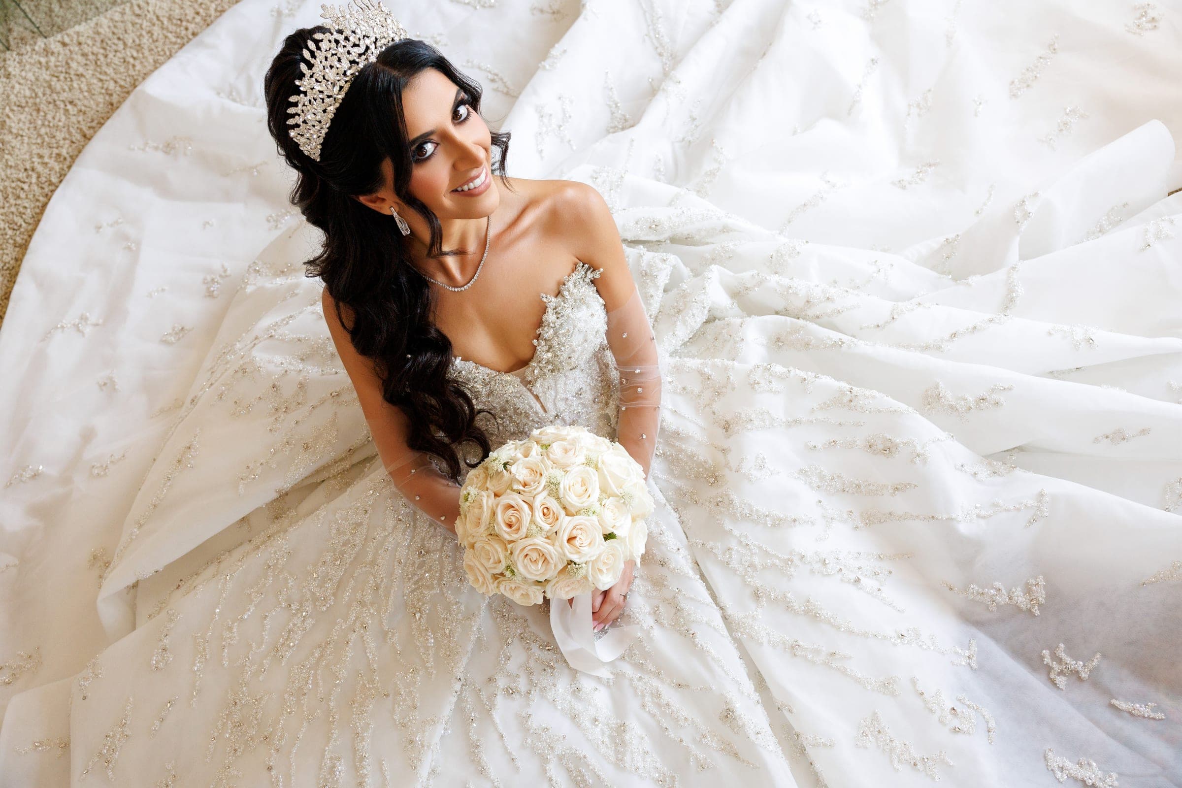 Bride seated in sparkling gown and crown holds pale roses, gazing upward with skirt spread around her.