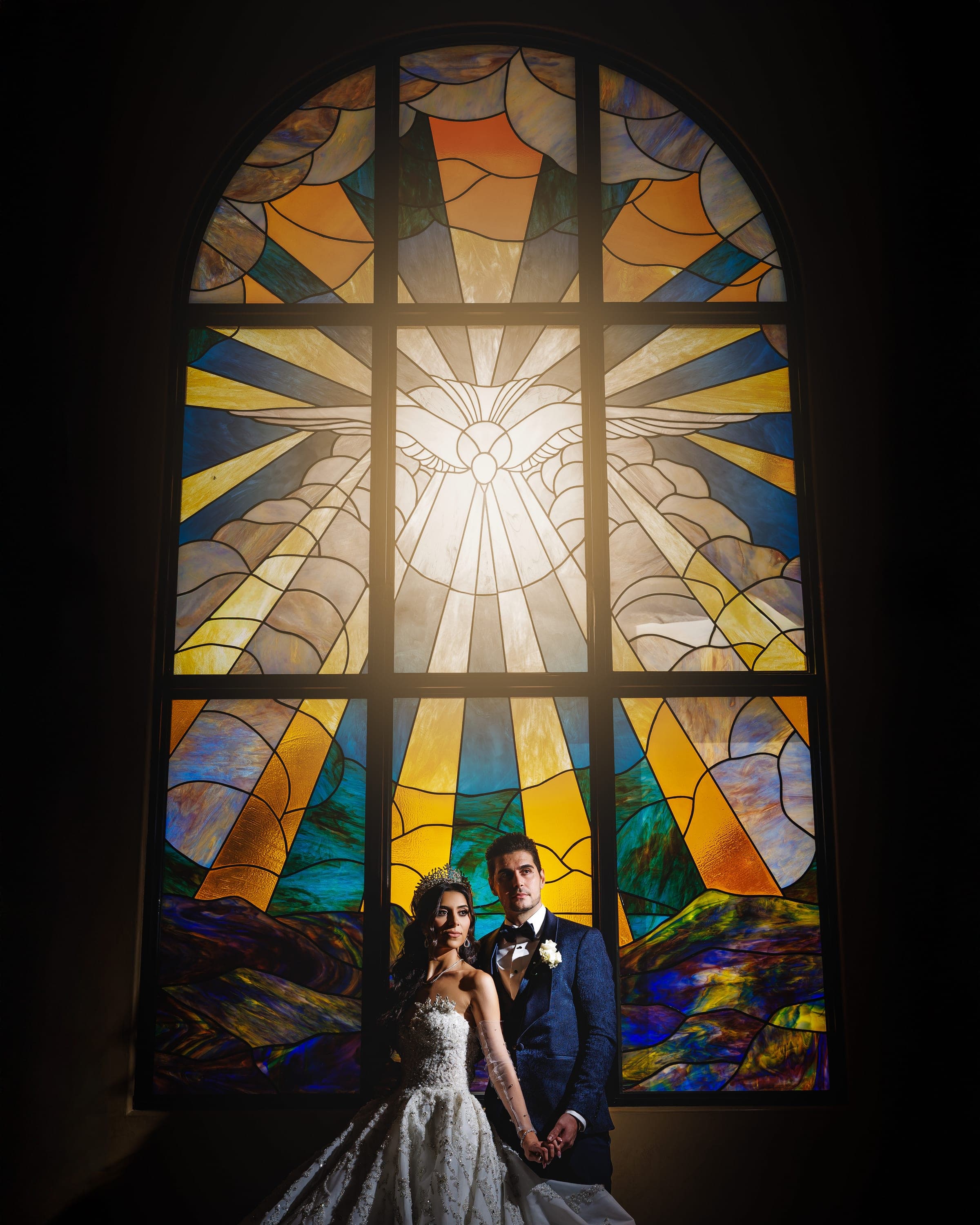 Bride and groom hold hands before a vast, glowing stained glass window with radiant colored beams.