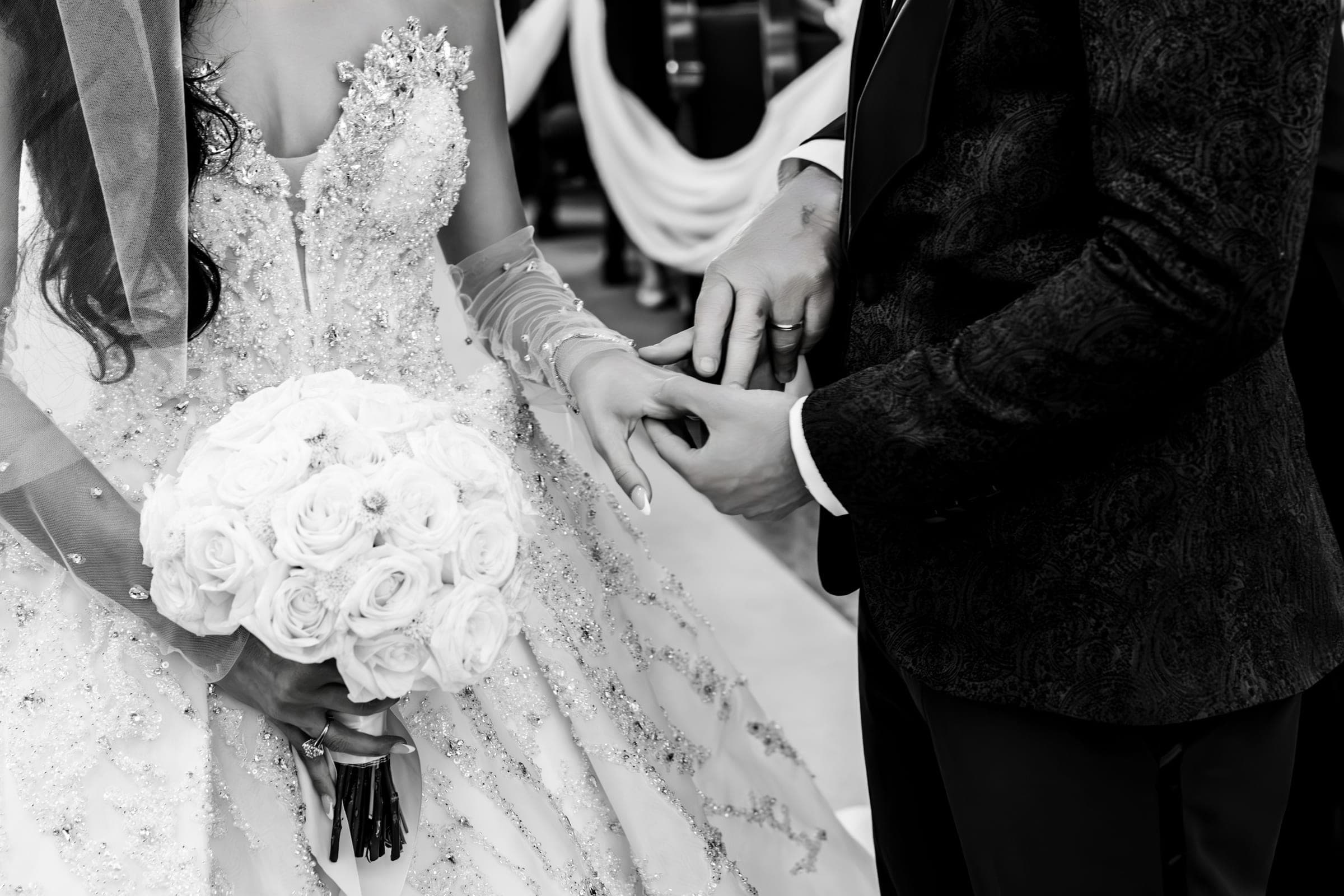 Black and white close-up of the couple holding hands, bride clutching a white rose bouquet in beaded gown