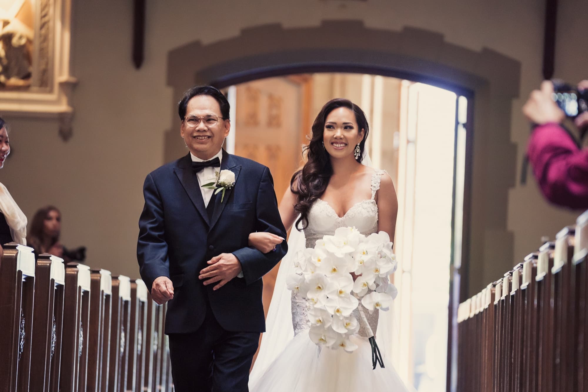 Caroline's father walking her down the church aisle, her cascading white orchid bouquet against the lace gown