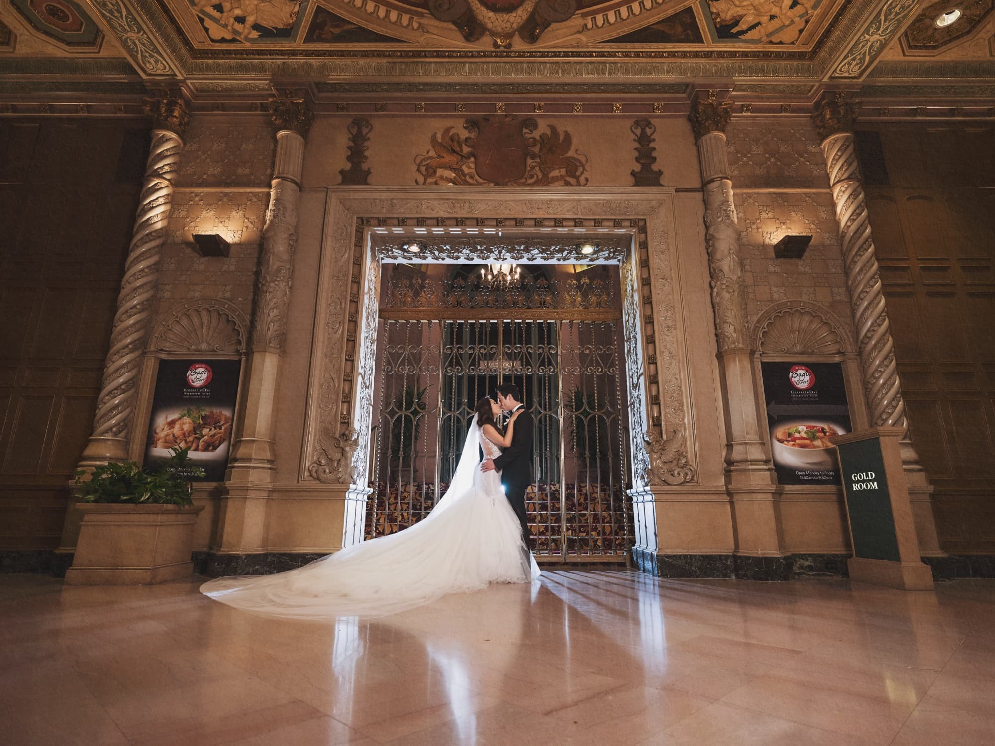 Caroline and Gerald kissing beneath the gilded entrance arch of the Millennium Biltmore with her cathedral veil pooling across the marble floor