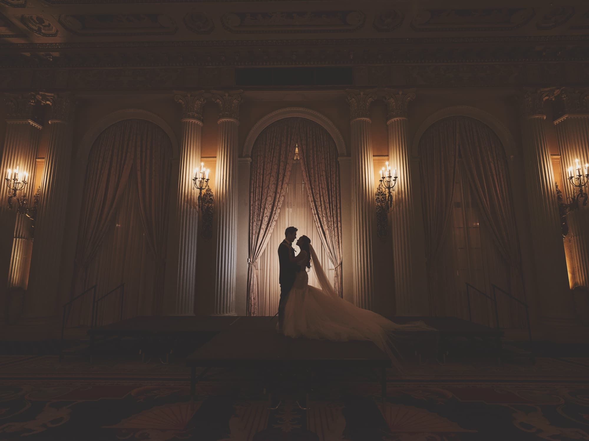 Silhouette of Caroline and Gerald between the draped arches and candlelit sconces of the Millennium Biltmore ballroom