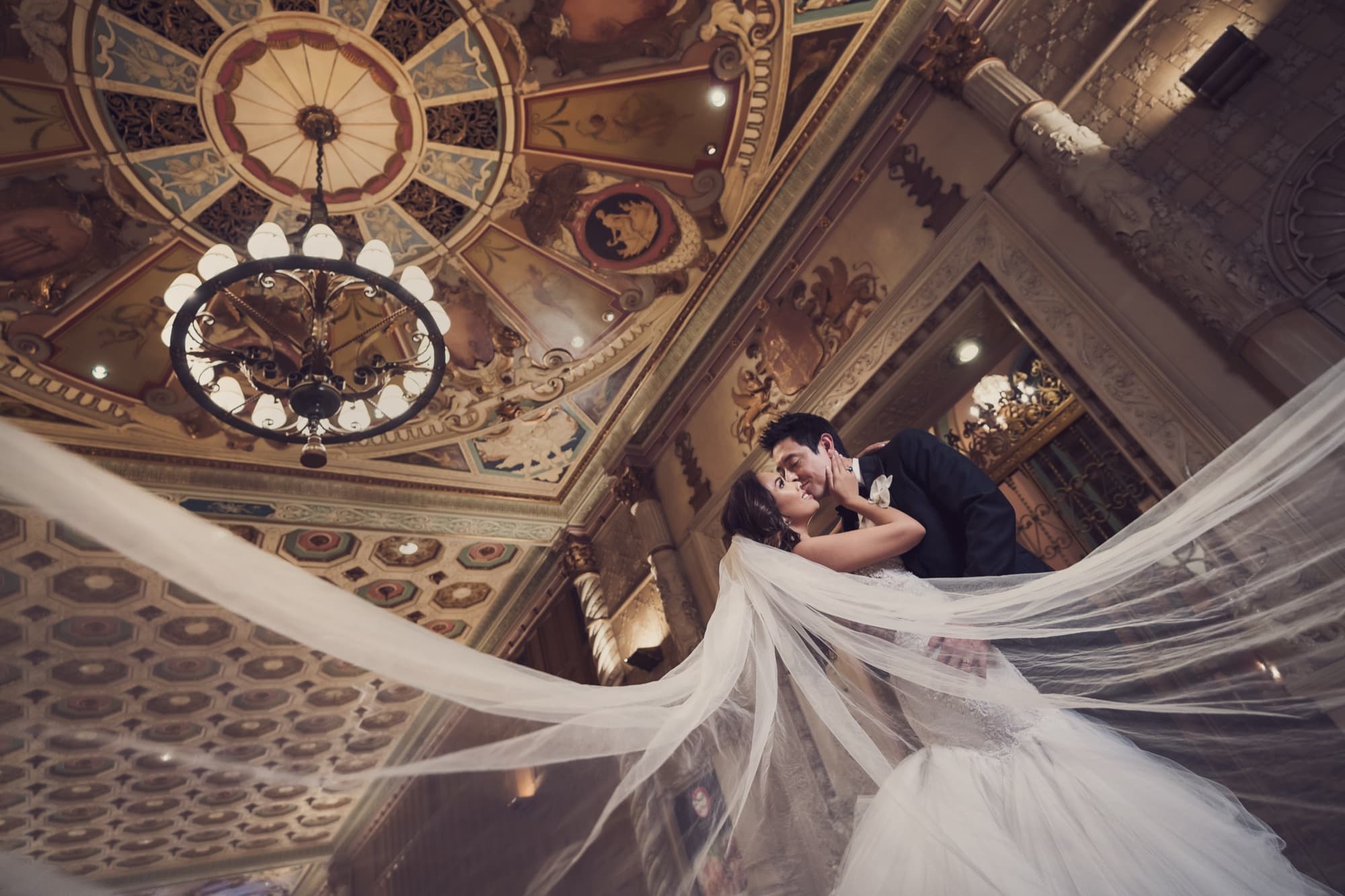 Caroline and Gerald embracing beneath the hand-painted ceiling of the Millennium Biltmore Los Angeles as her cathedral veil sweeps through the frame, photographed by Michael Anthony Photography