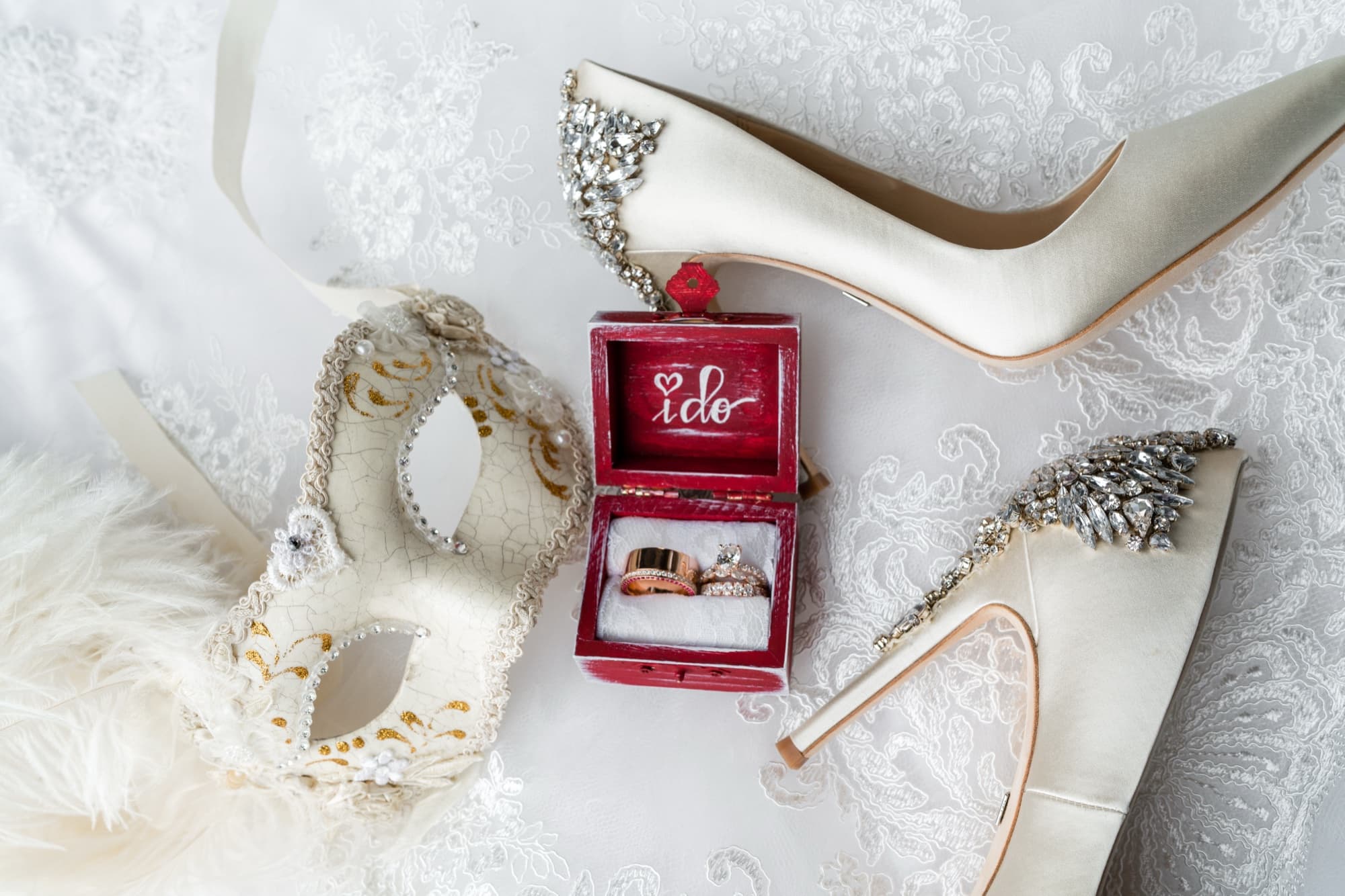 Masquerade flat lay with a feathered Venetian mask, crystal-buckled bridal heels, and a red I do ring box holding gold bands