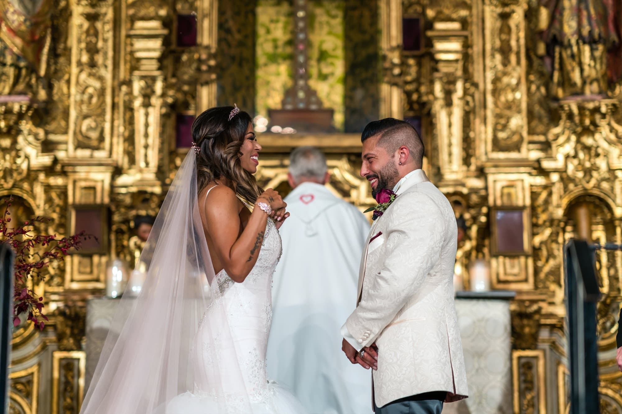 Bride and groom laughing together during vows in front of the gilded gold altar of the chapel at the Mission Inn