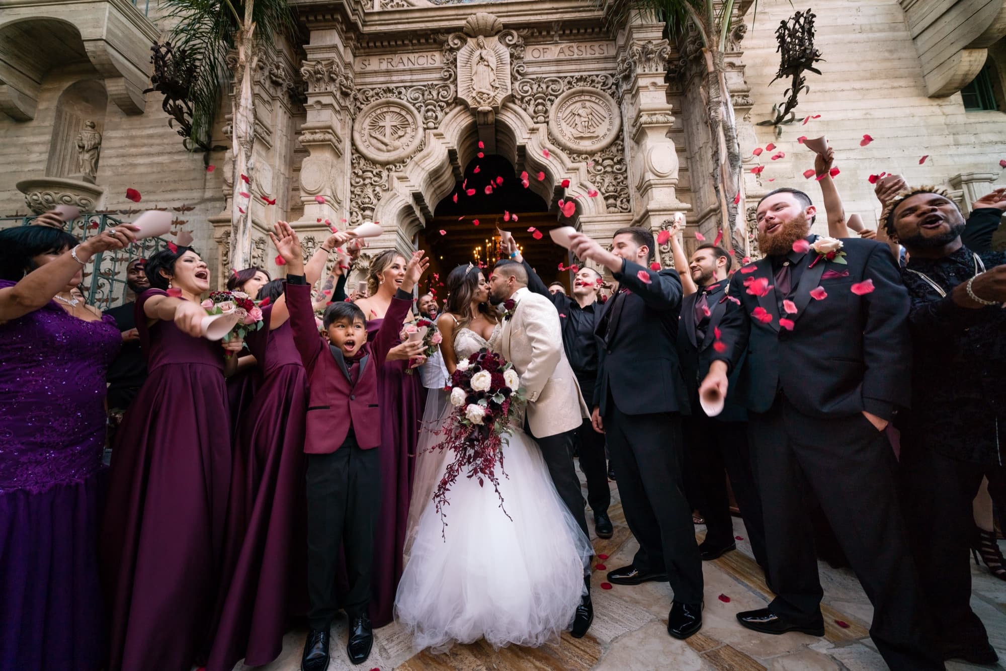 Wedding party in plum and black throwing rose petals over the bride and groom as they kiss outside the Mission Inn chapel