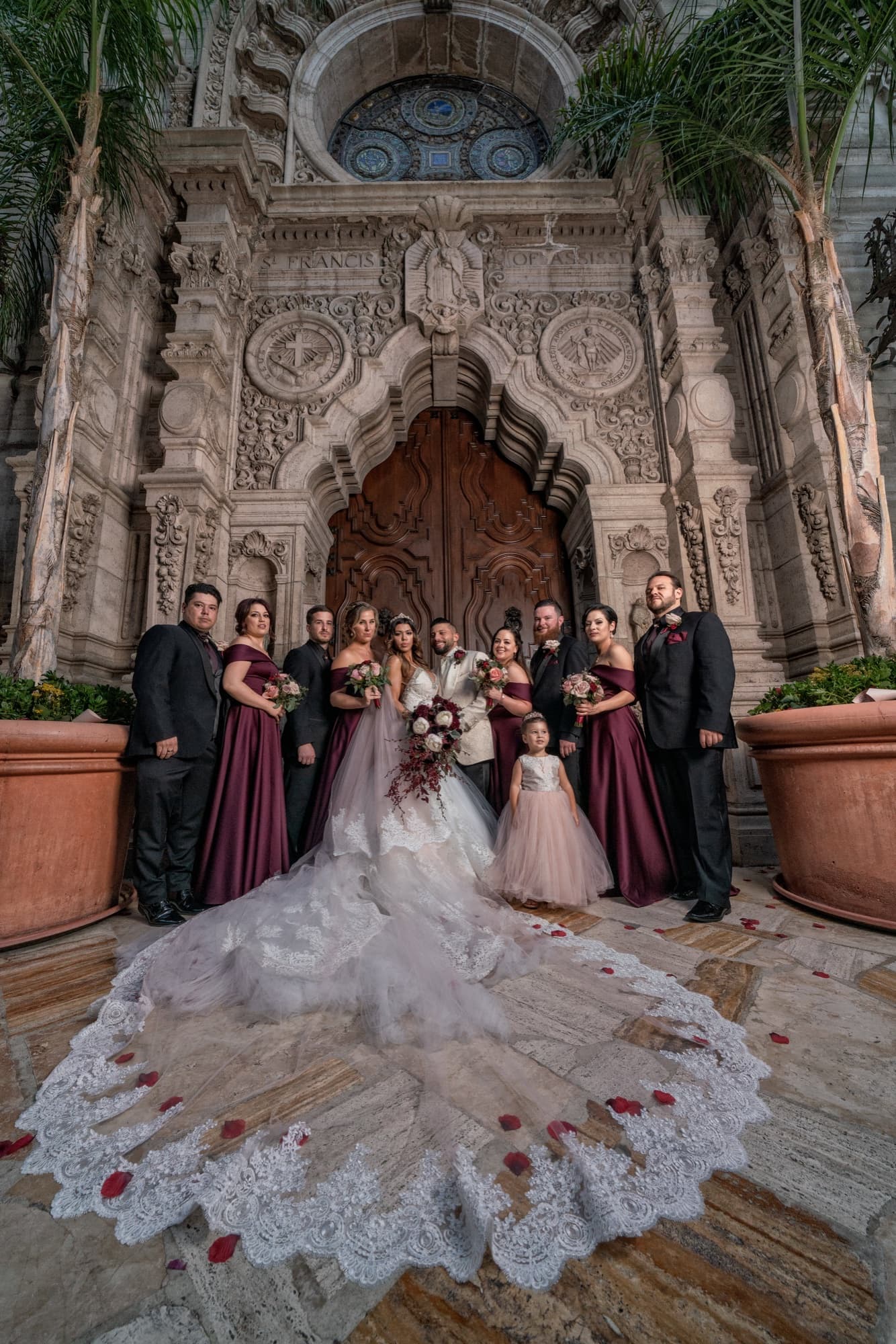Full wedding party in plum and burgundy surrounding the bride, her lace train covering the stone steps of the Mission Inn entrance