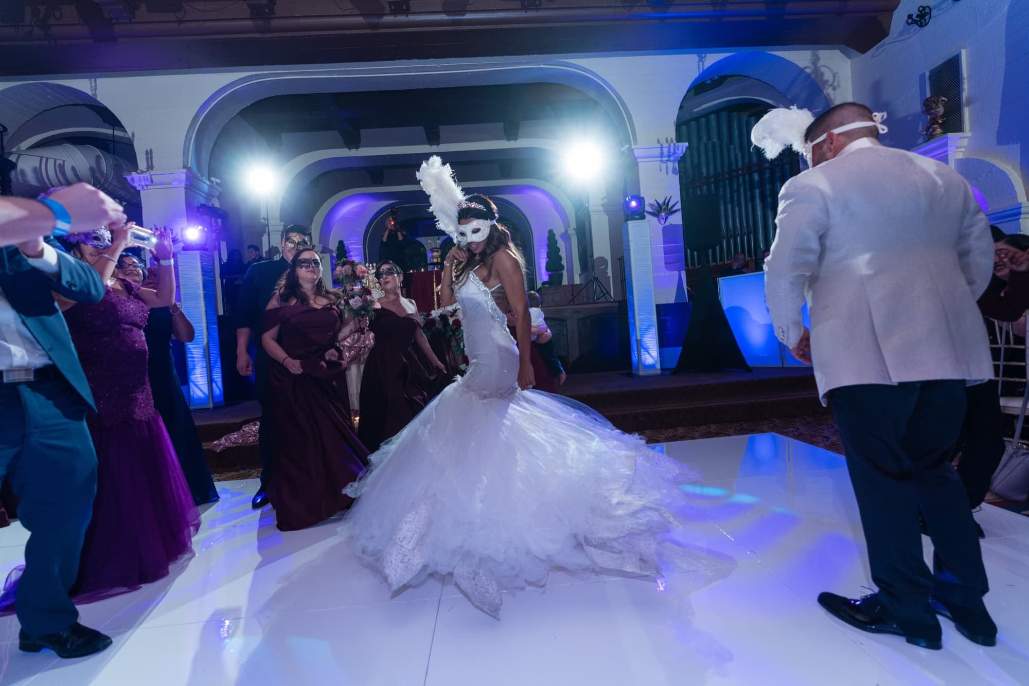 Bride in a feathered masquerade mask dancing with the wedding party on the white dance floor at the Mission Inn reception