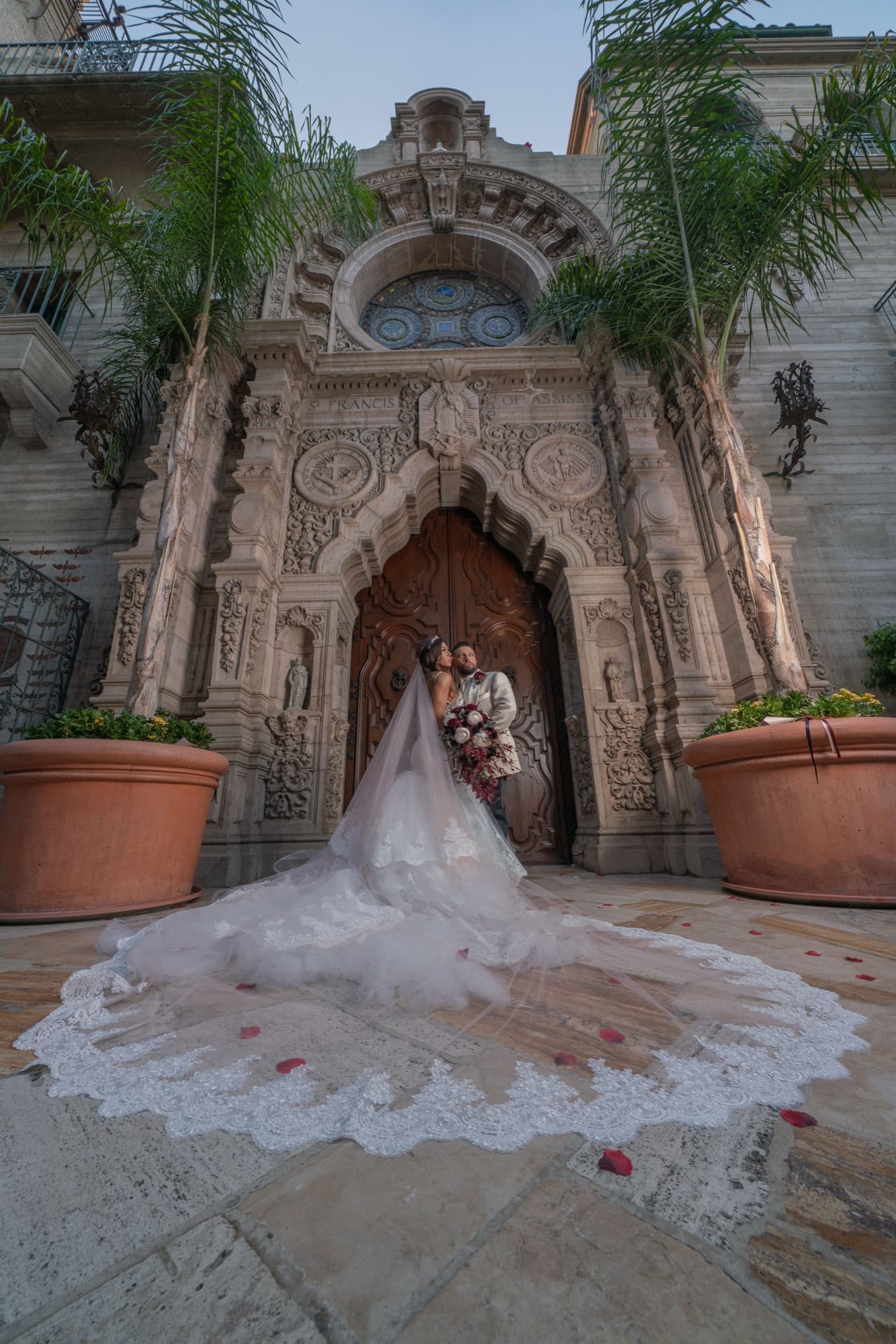 Bride and groom framed by the carved stone entrance of the Mission Inn with her lace cathedral train spread across the steps, photographed by Michael Anthony Photography