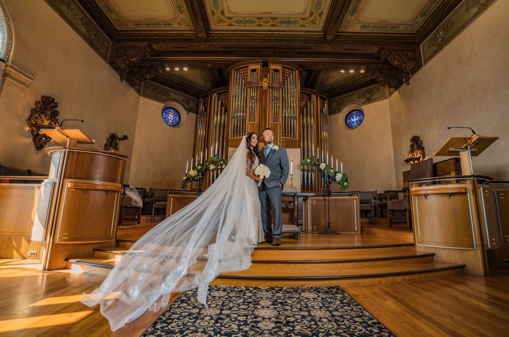 Karina and Faustino on the altar steps of The Neighborhood Church beneath the wooden pipe organ, her veil cascading down the steps