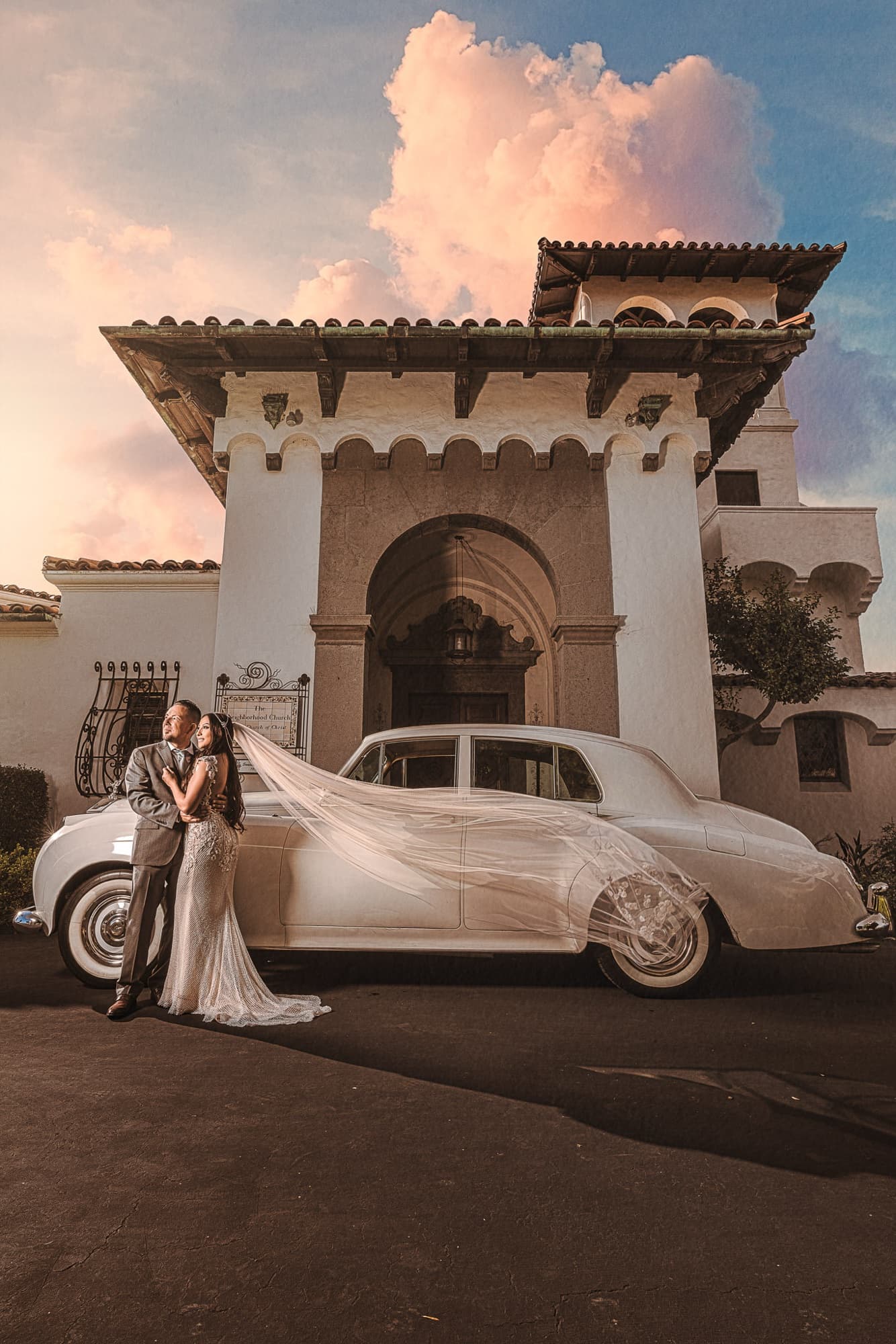 Karina and Faustino embracing beside a classic white car in front of a Mediterranean stone tower at sunset in Palos Verdes
