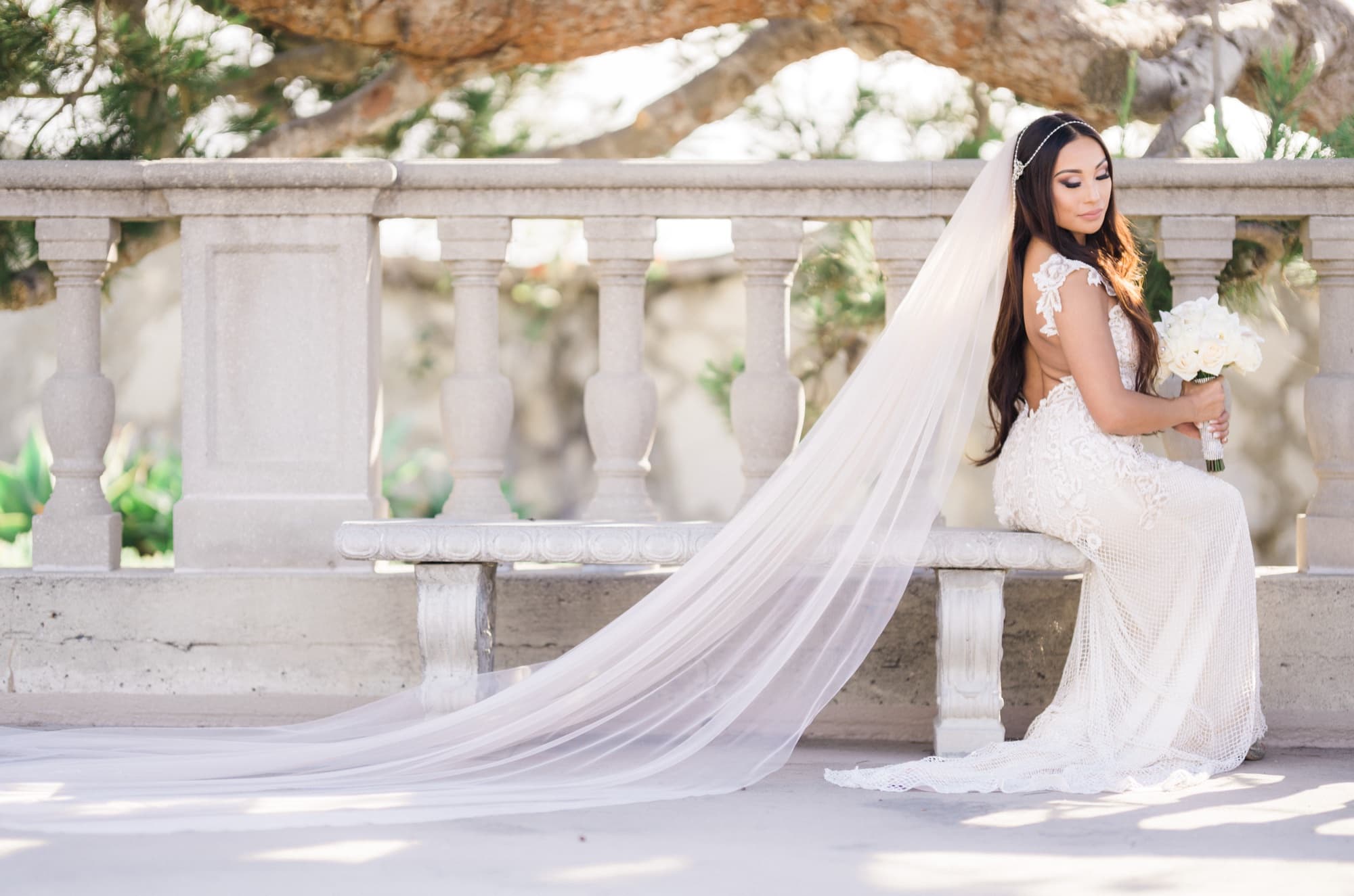 Karina seated on a stone balustrade bench with her cathedral veil cascading across the ground in Palos Verdes