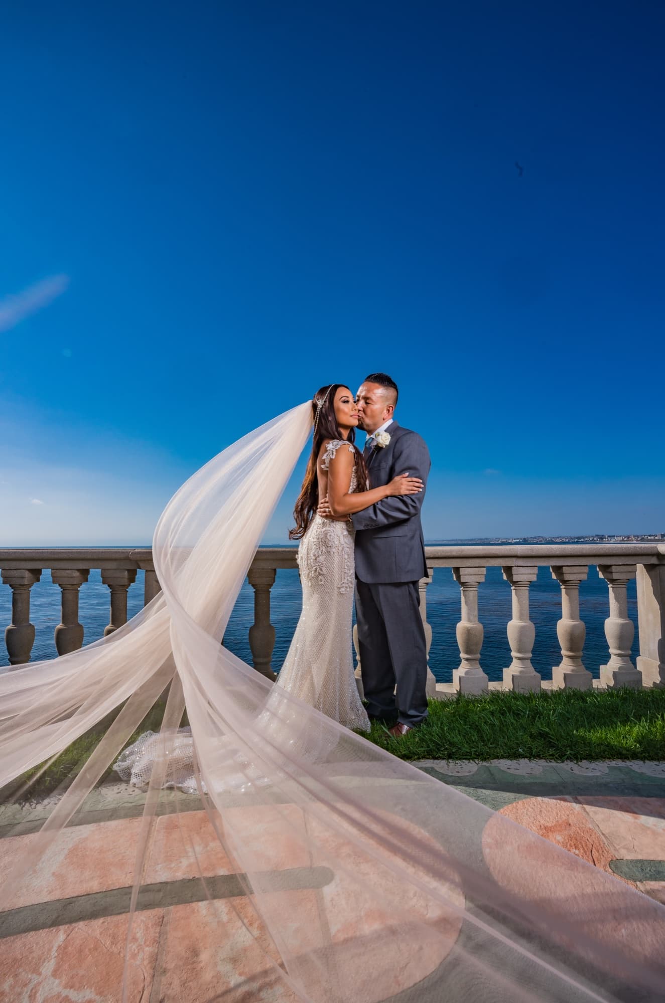 Karina and Faustino embracing on the ocean-view balustrade in Palos Verdes at dusk, her cathedral veil streaming across the stone under a deep blue sky, photographed by Michael Anthony Photography