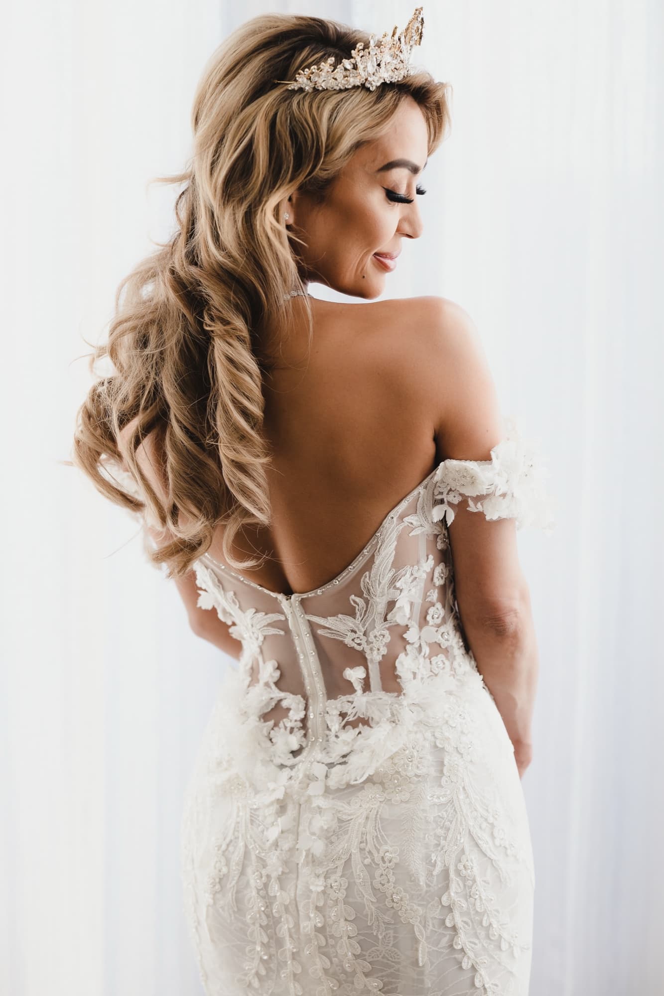 Danielle from behind in her illusion-back lace gown, long curled hair over one shoulder and crown in place