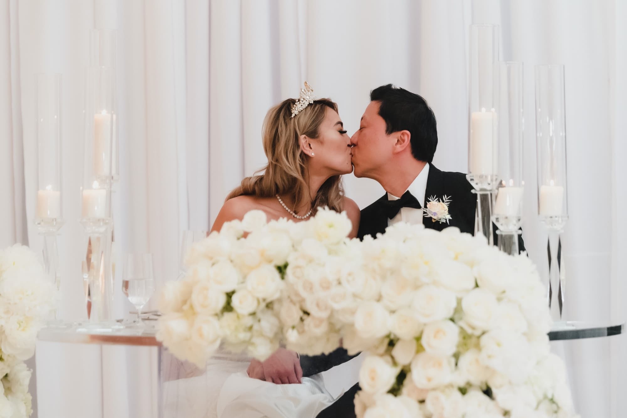 Danielle and Justin kissing at the sweetheart table behind a bank of white roses and candlelight