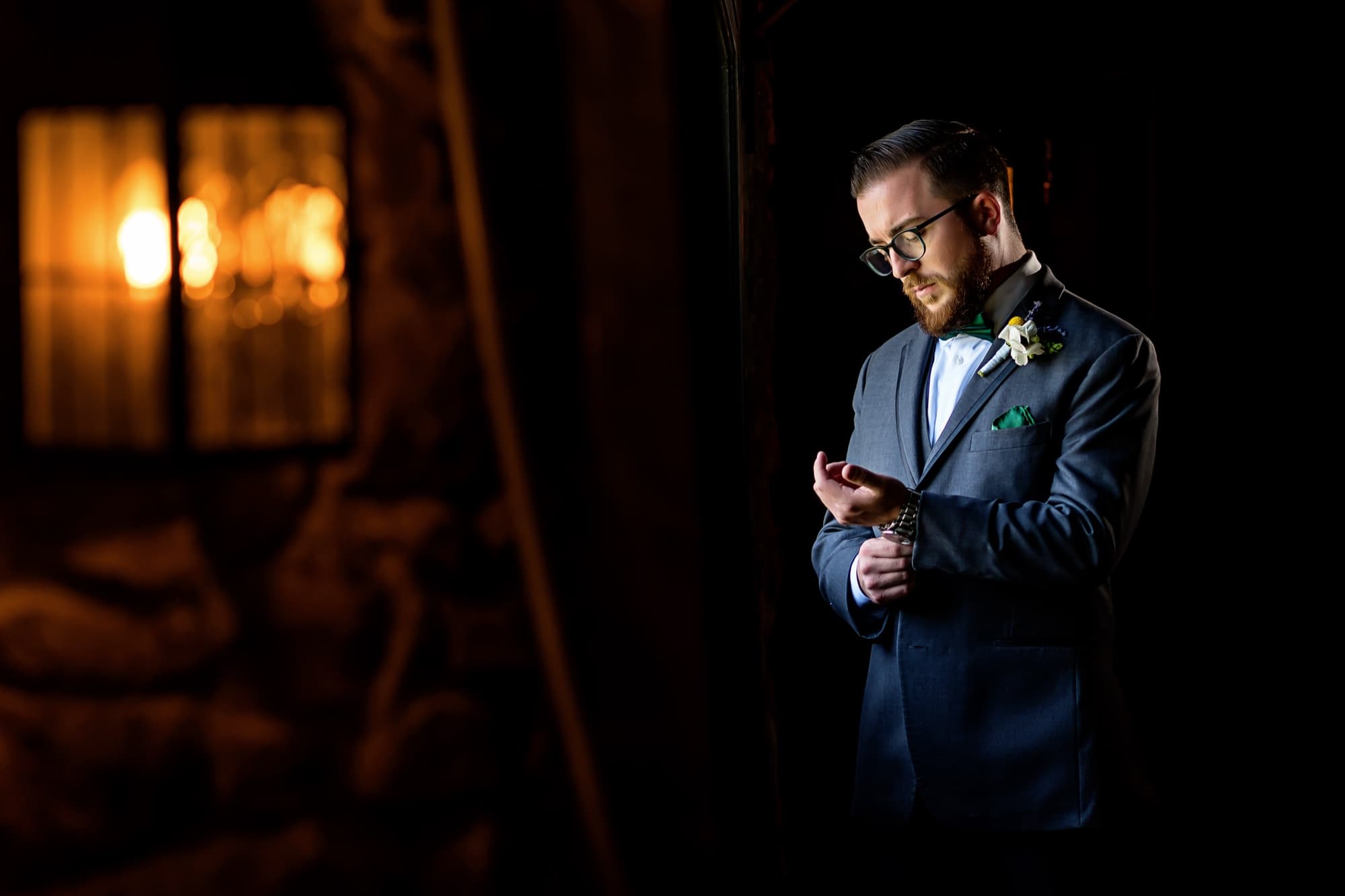 Adam in a gray suit and emerald bowtie lit by lantern light in the stone entry of Newhall Mansion