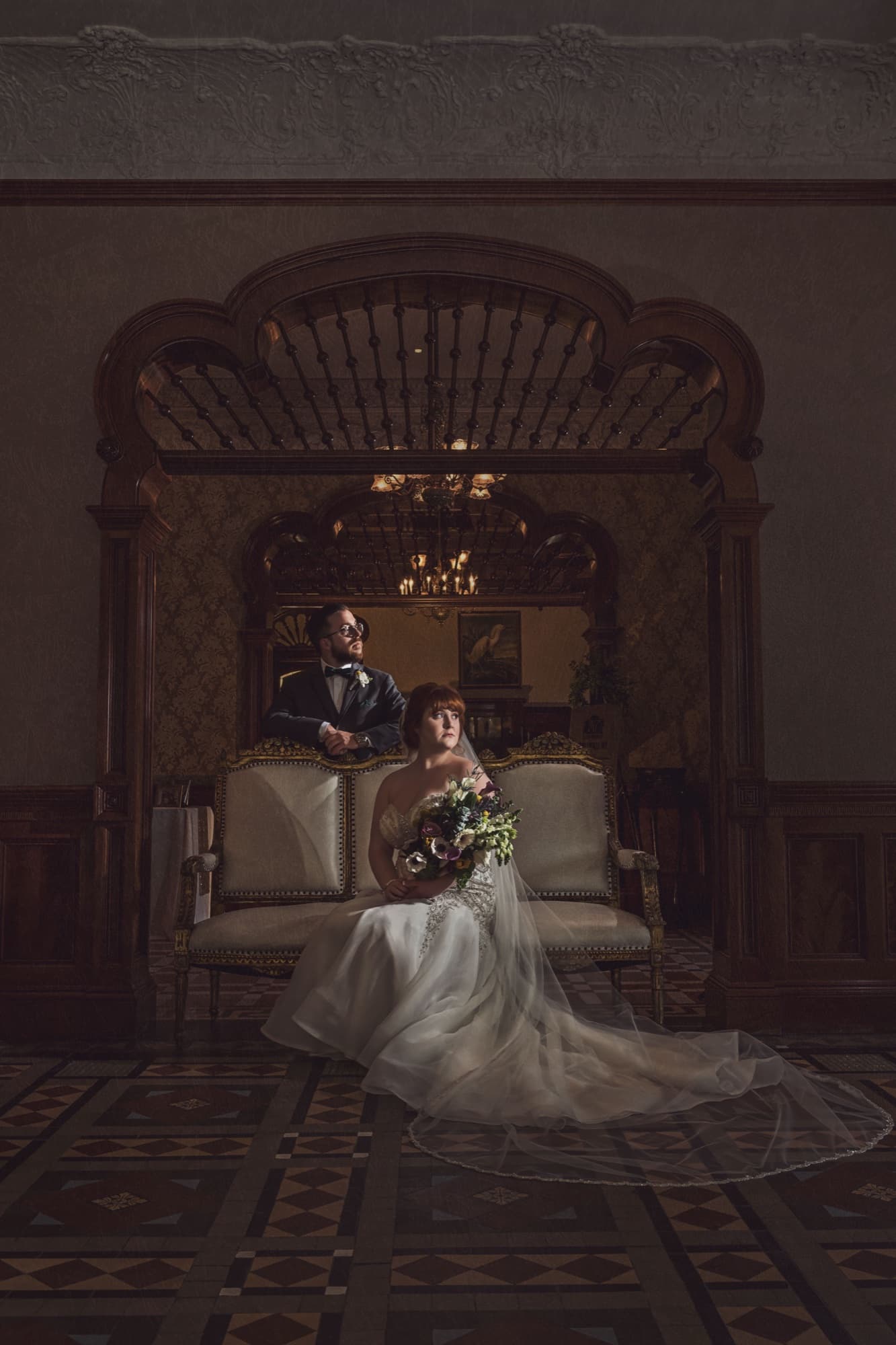 Michelle seated in a wingback chair in the mansion's dark-paneled parlor, her gown and bouquet glowing in the low light