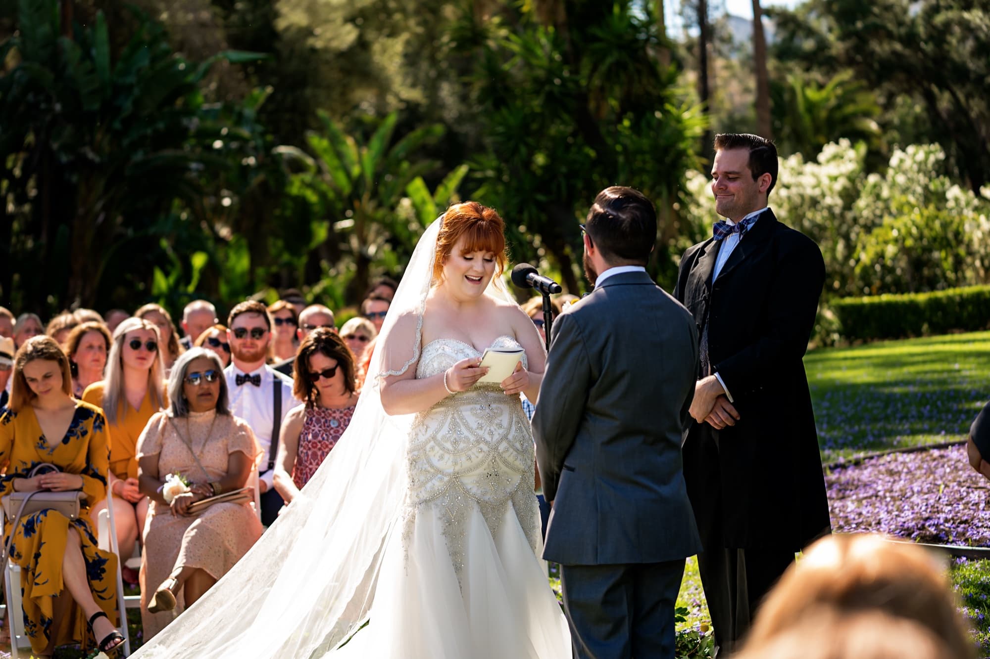 Michelle reading her vows to Adam under the garden trees at Newhall Mansion as guests look on
