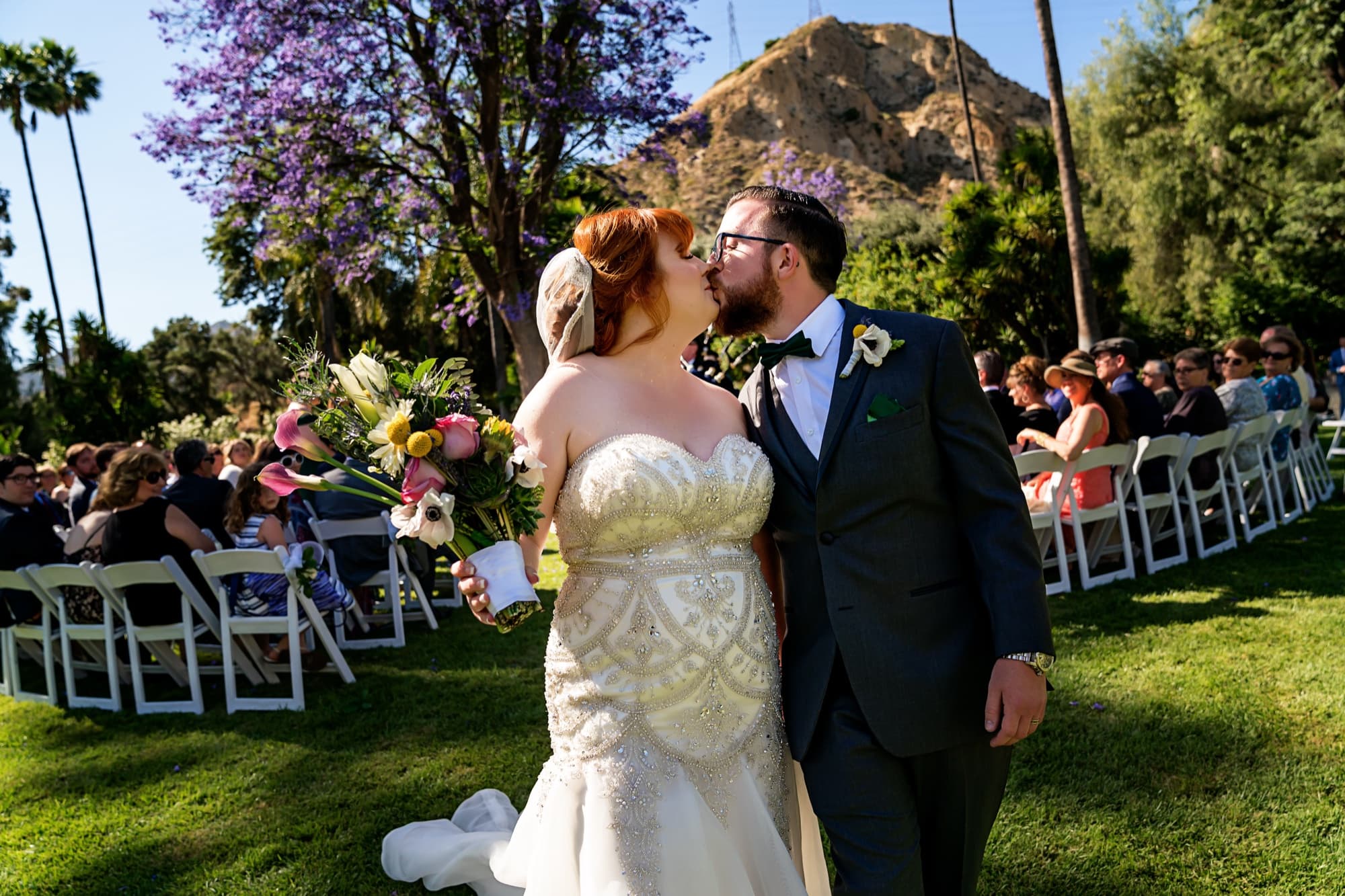 Michelle and Adam kissing at the end of the aisle with the jacaranda blooming and the Piru hillside behind them