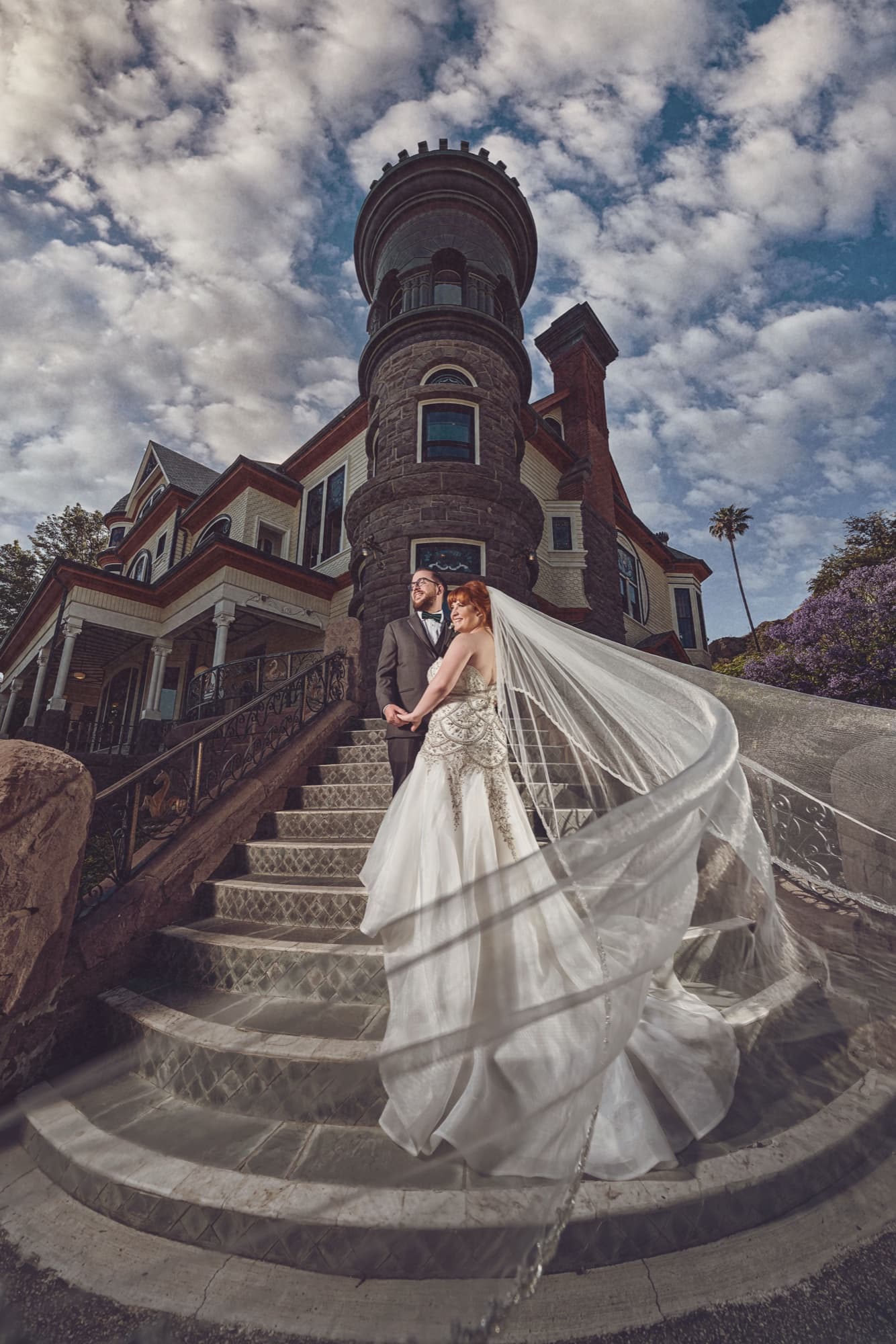 Michelle and Adam on the turret steps of Newhall Mansion beneath dramatic clouds, her veil sweeping down the stone