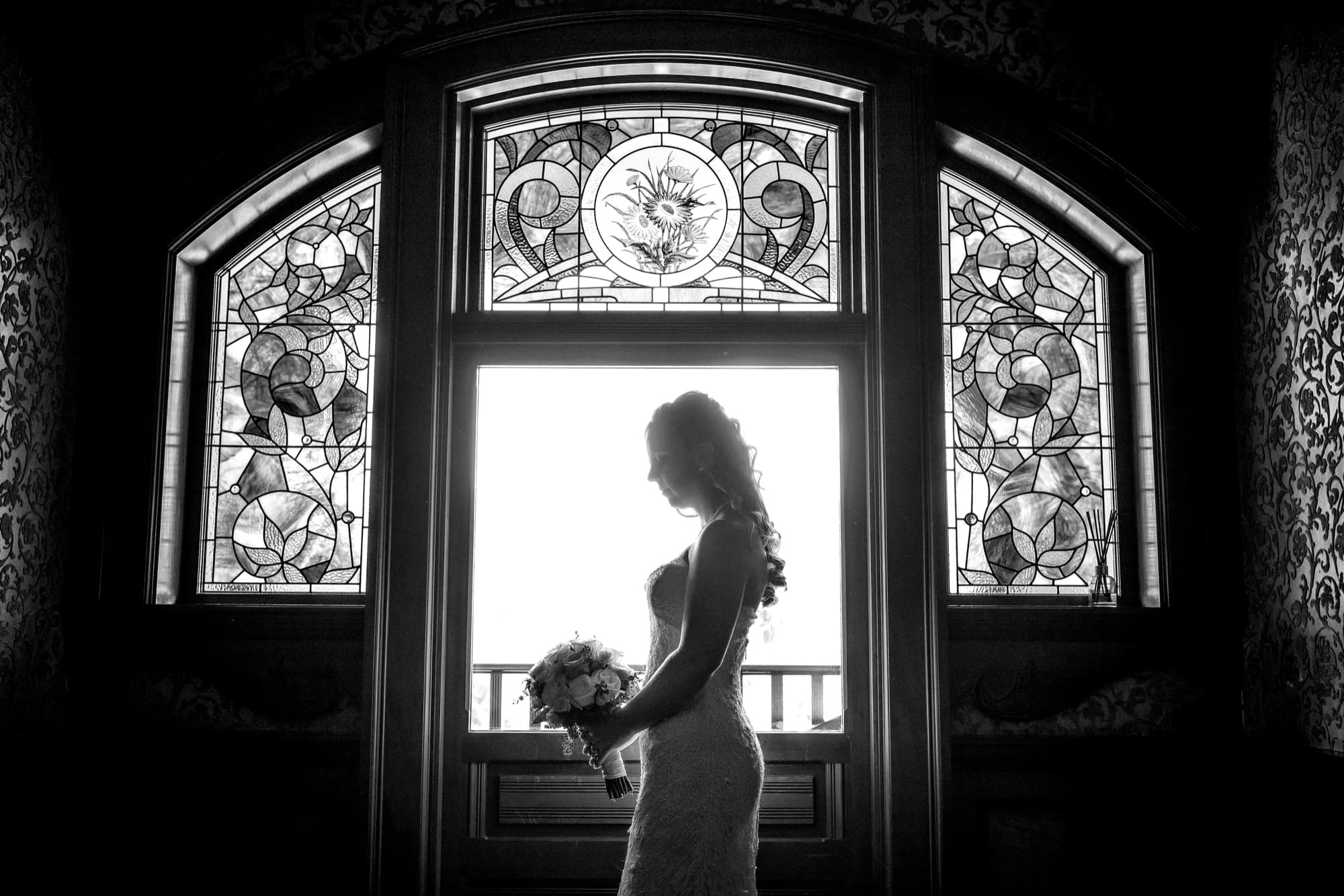 Black and white silhouette of Cathryn holding her bouquet in the stained-glass landing window at Newhall Mansion