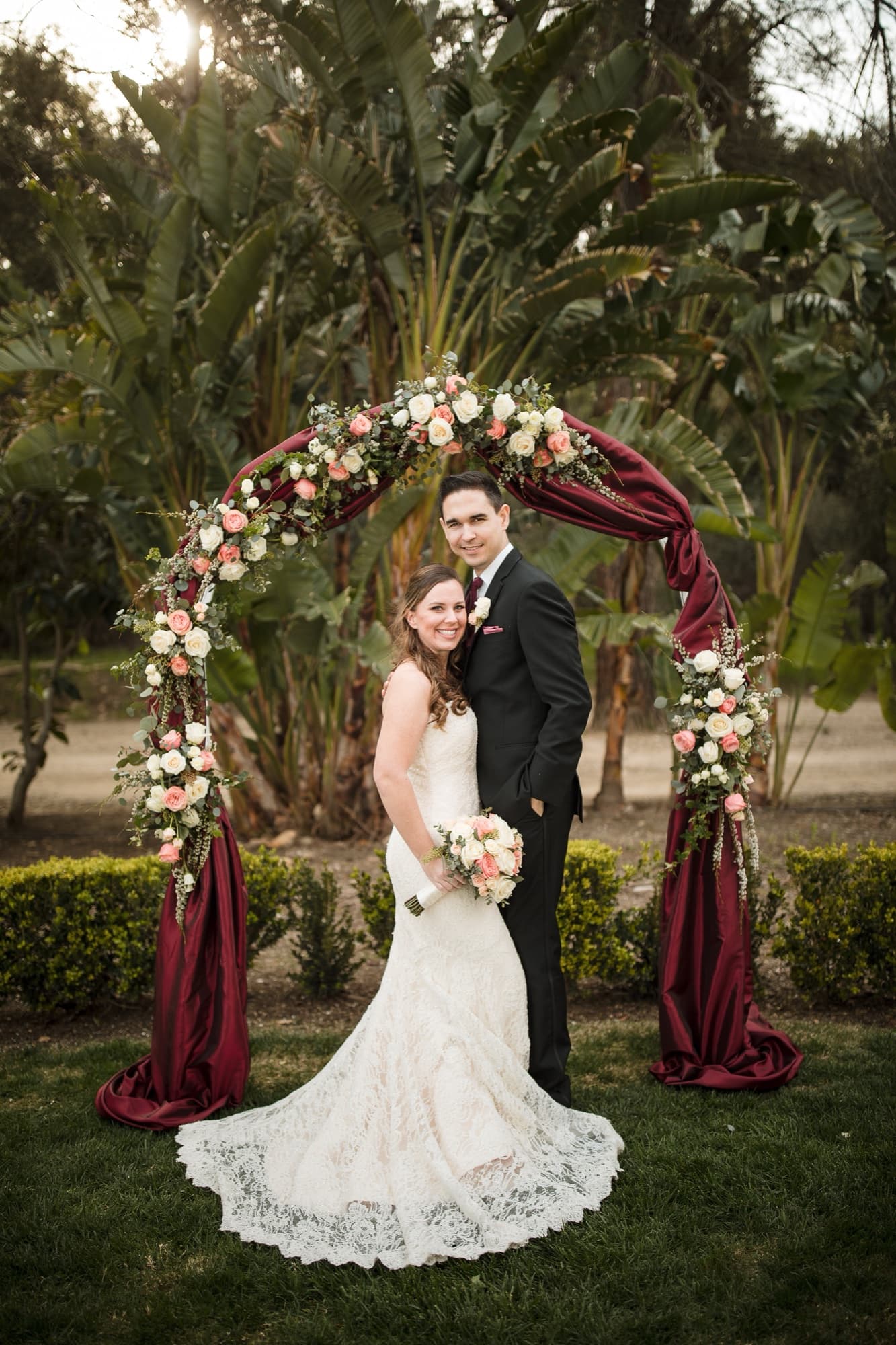 Cathryn and Tommy beneath the circular blush rose arch wrapped in burgundy drape in the Newhall Mansion garden