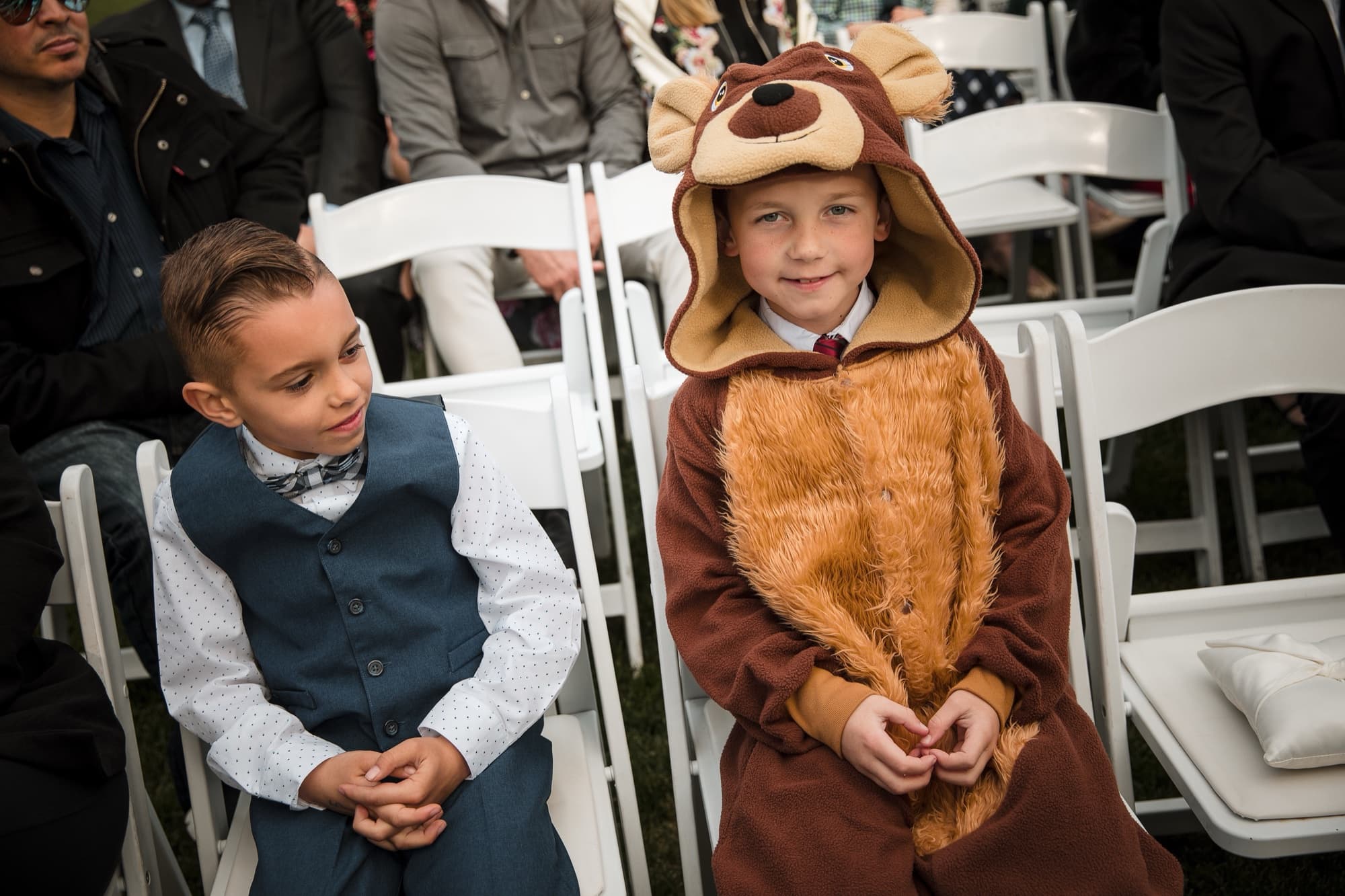 The ring bearer in a full dog costume sitting beside his brother in the ceremony chairs, both waiting patiently
