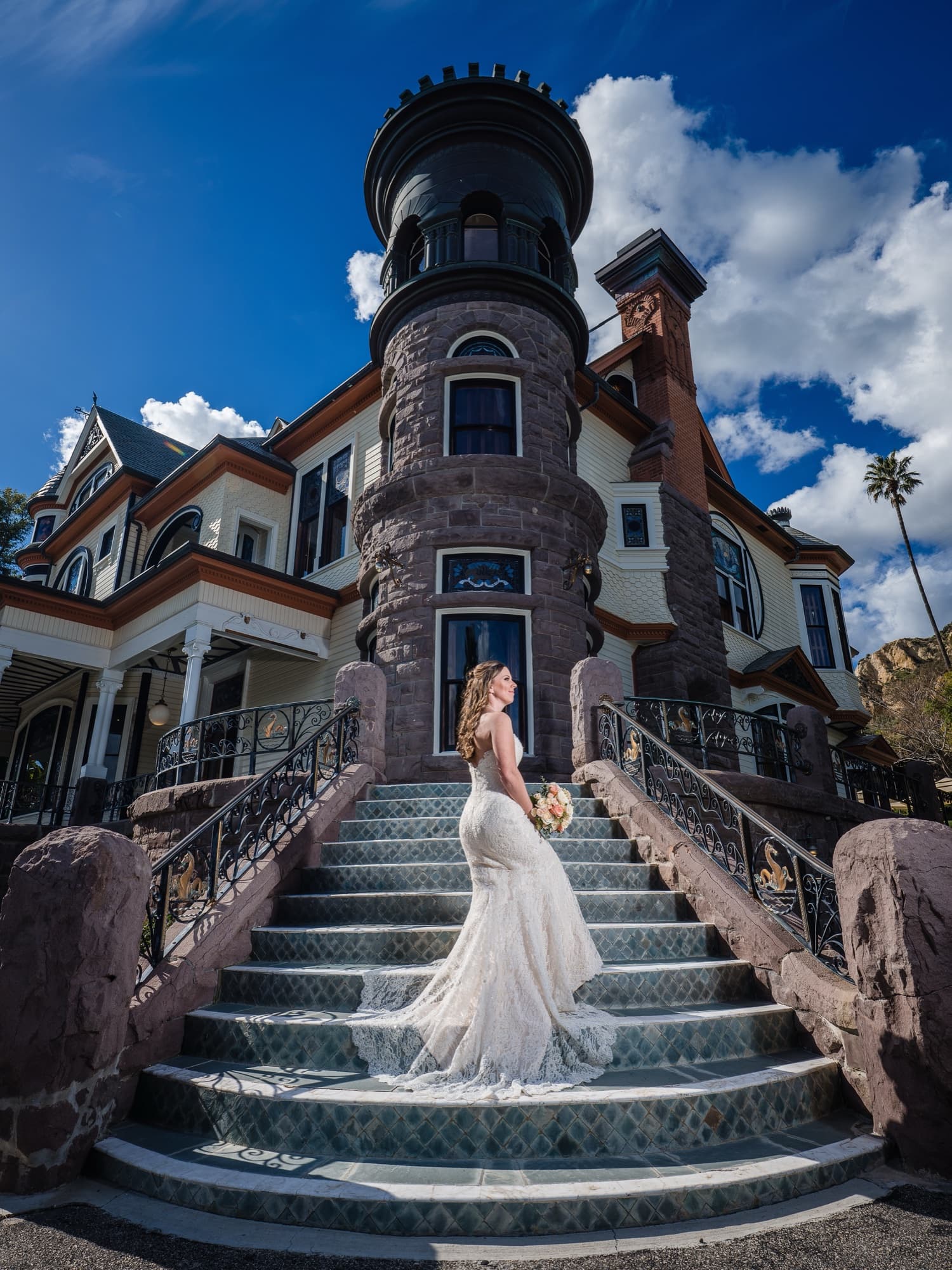 Cathryn in her lace gown on the tiled staircase beneath the stone turret of Newhall Mansion under a dramatic blue sky, photographed by Michael Anthony Photography