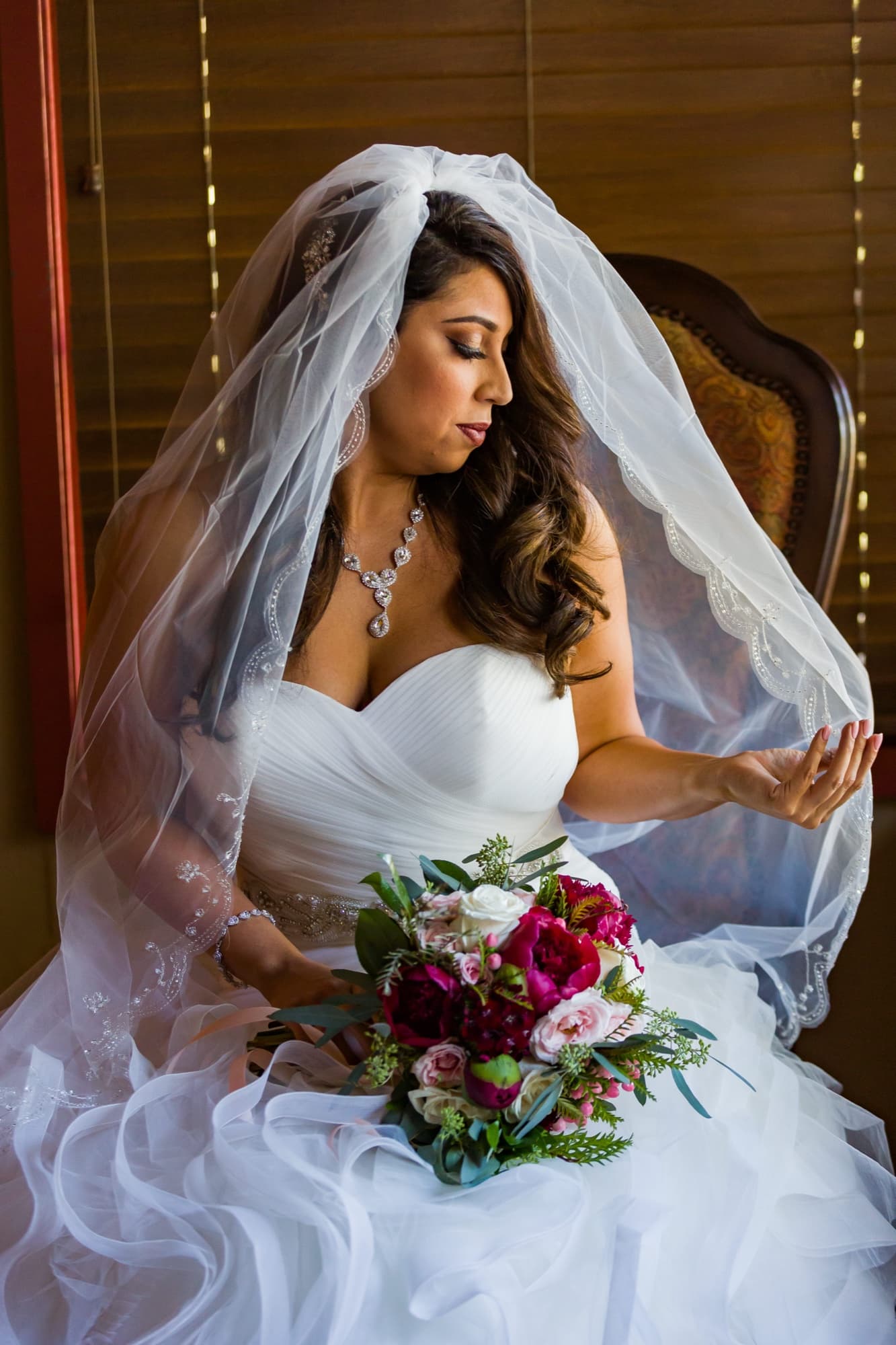 Soraya seated in the window light at Padua Hills Theatre, cathedral veil over her shoulders and a deep red bouquet in her lap