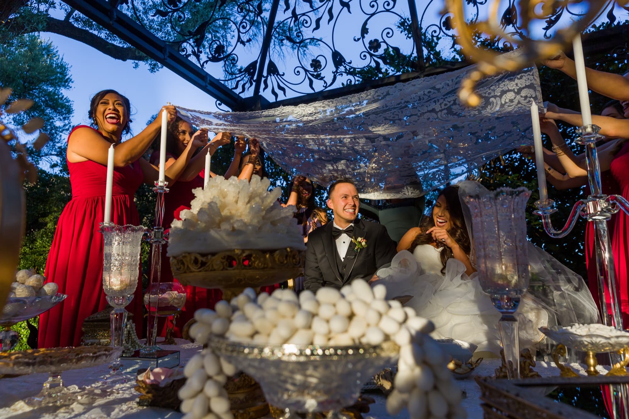 Family grinding sugar cones over the lace canopy held above Soraya and Donald during the sugar ceremony at their Persian wedding