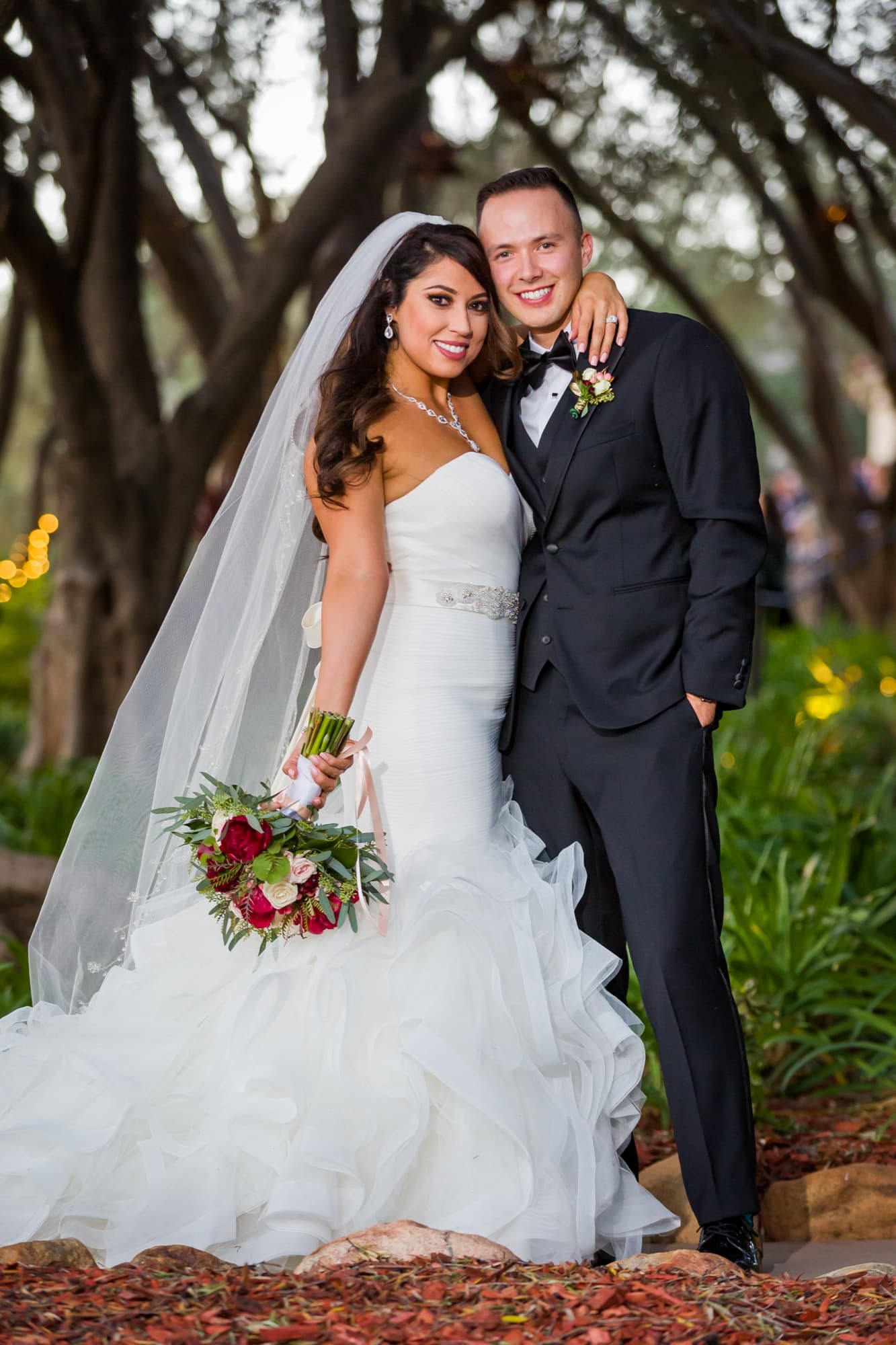 Soraya and Donald standing together in the olive grove at Padua Hills Theatre with her deep red bouquet and cathedral veil