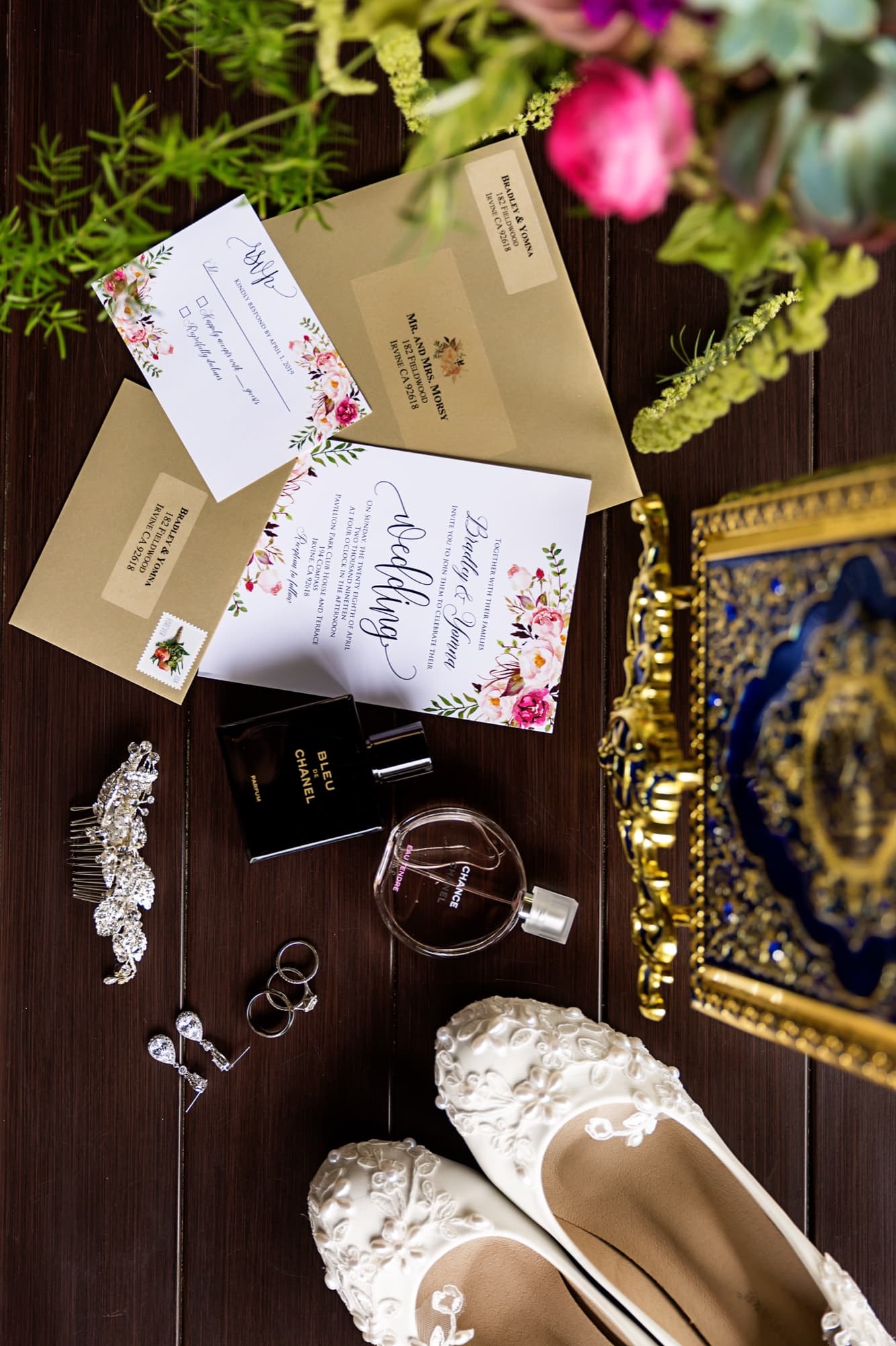 Wedding flat lay with the invitation suite, lace heels, rings, perfume, and an ornately gilded heirloom book on dark wood