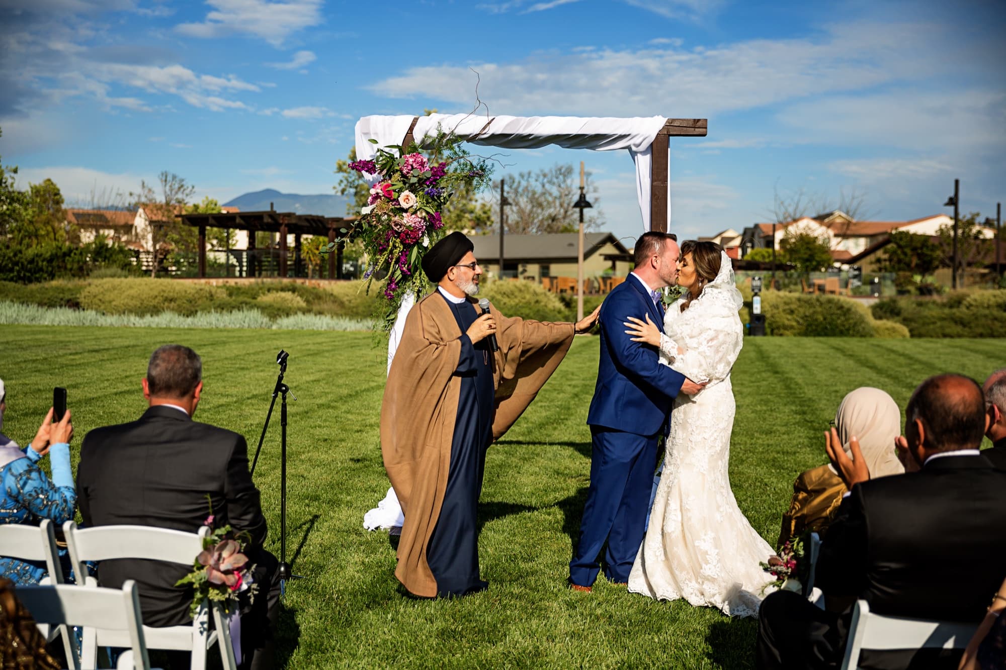 Yomna and Bradley kissing beneath the floral-dressed wooden arbor as the officiant and guests look on at Pavilion Park