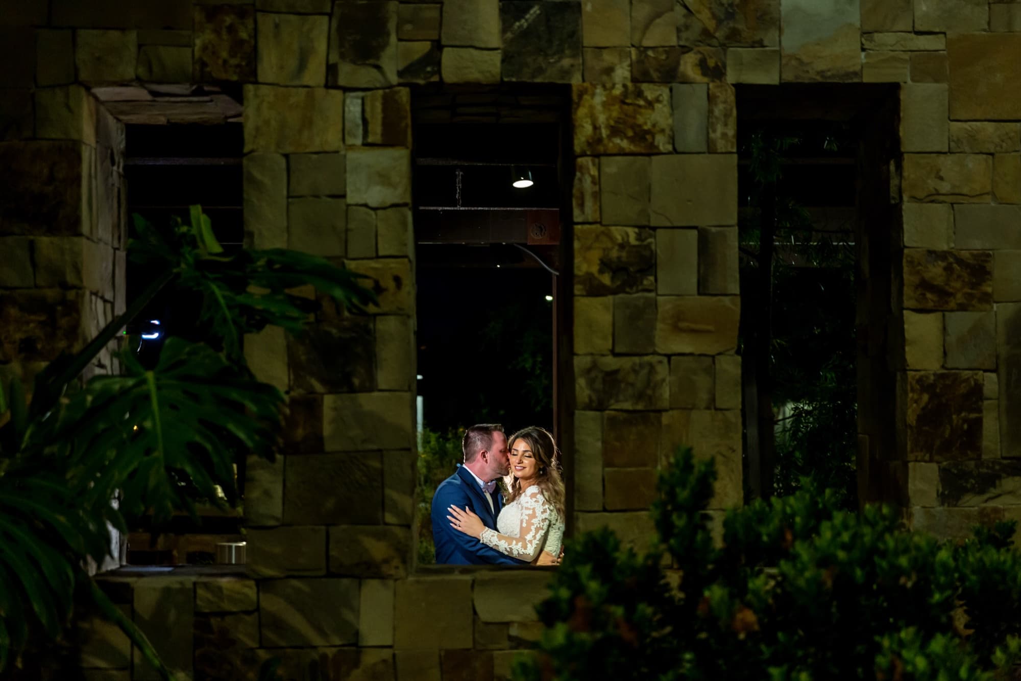 Yomna and Bradley embracing in a pool of light against the stone wall of the Pavilion Park clubhouse at night