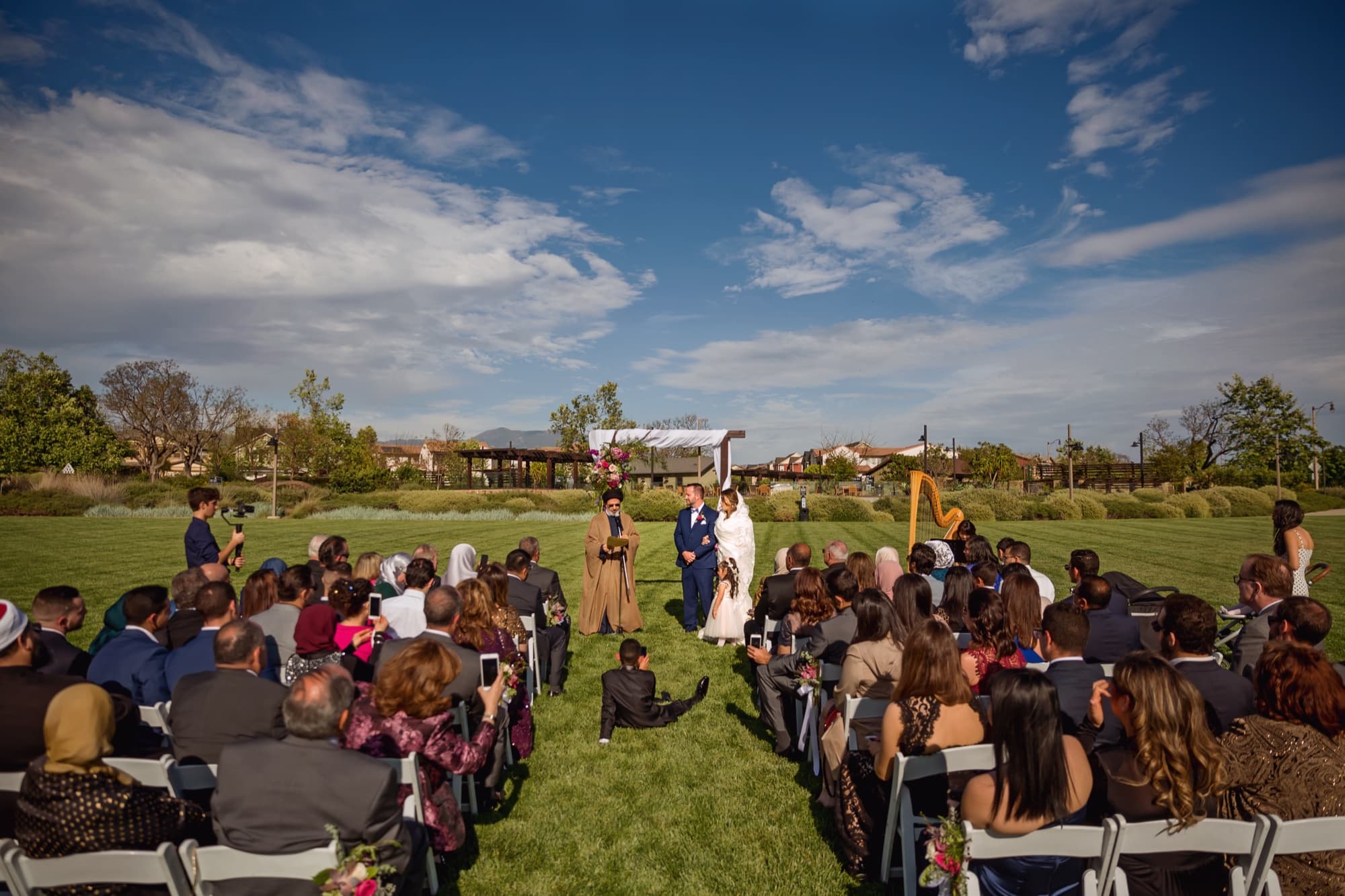 Wide view of Yomna and Bradley's outdoor wedding ceremony on the lawn at The Clubhouse at Pavilion Park in Irvine under a dramatic blue spring sky, photographed by Michael Anthony Photography