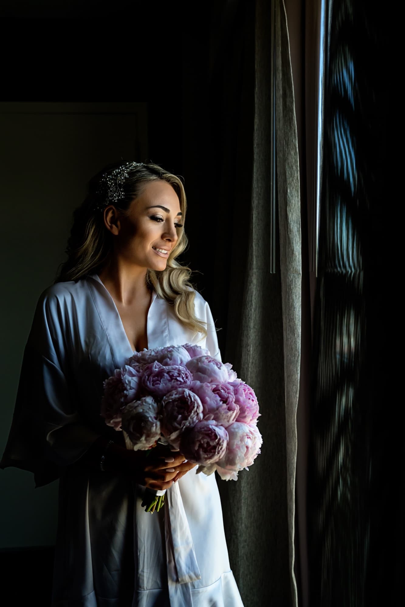 Laura in a silk robe holding an armful of blush peonies in the window light before her Saint Sophia Cathedral wedding