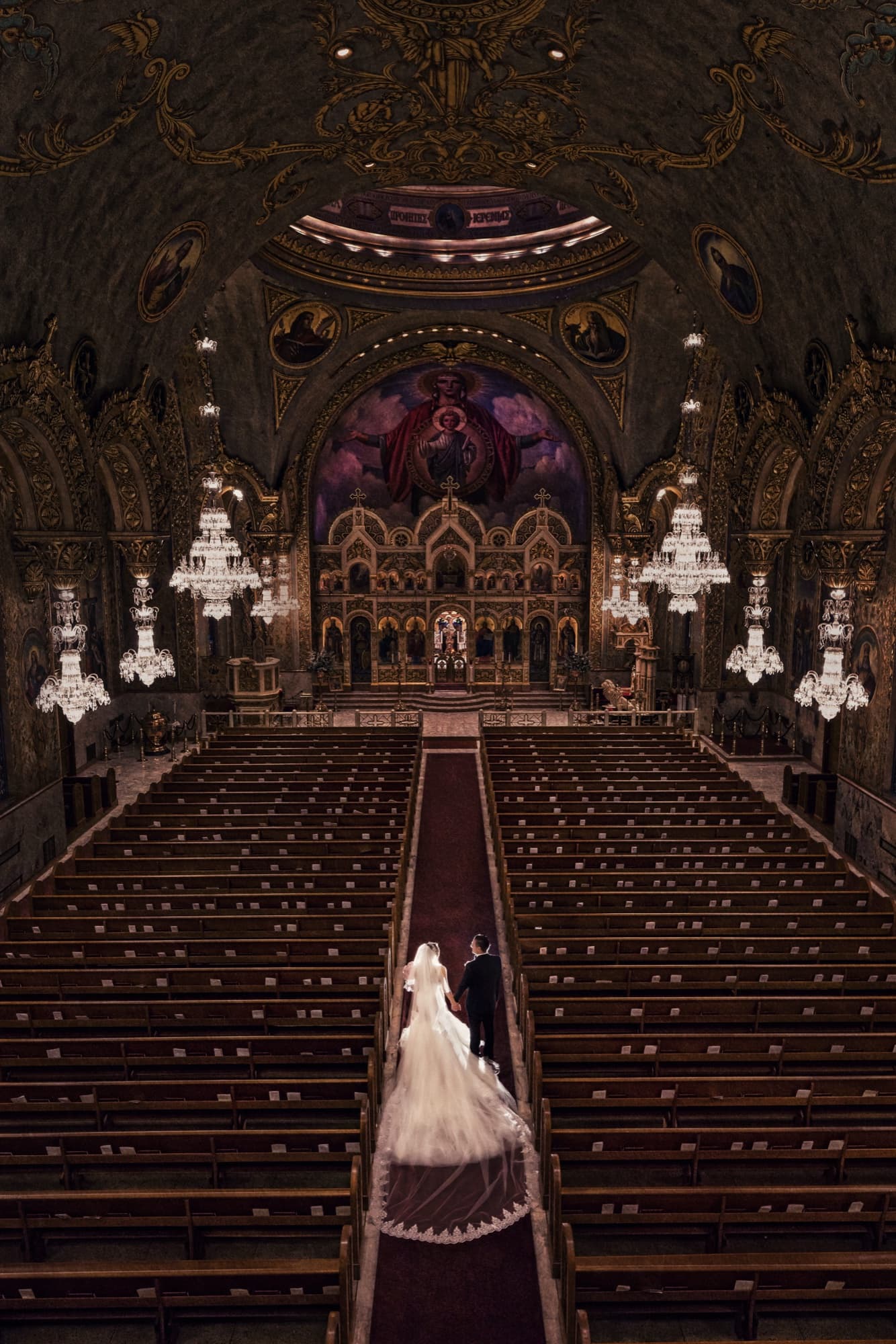 Laura and Garen standing small at the end of the aisle beneath the gilded domes and crystal chandeliers of Saint Sophia Cathedral in Los Angeles, photographed by Michael Anthony Photography