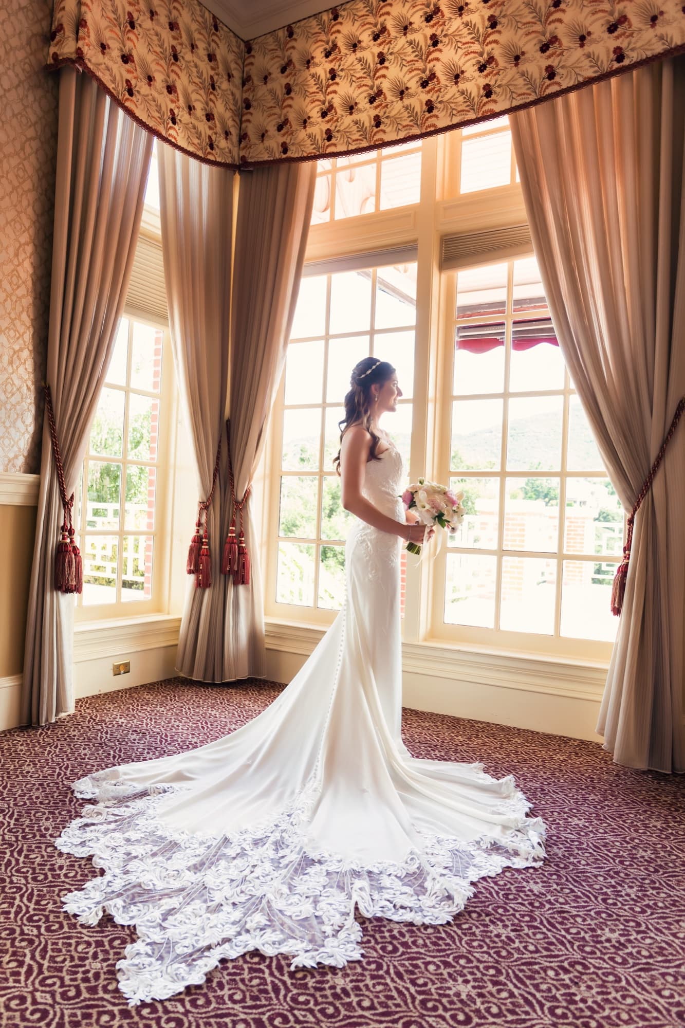Heather at the tall clubhouse windows with her lace train fanned across the patterned carpet