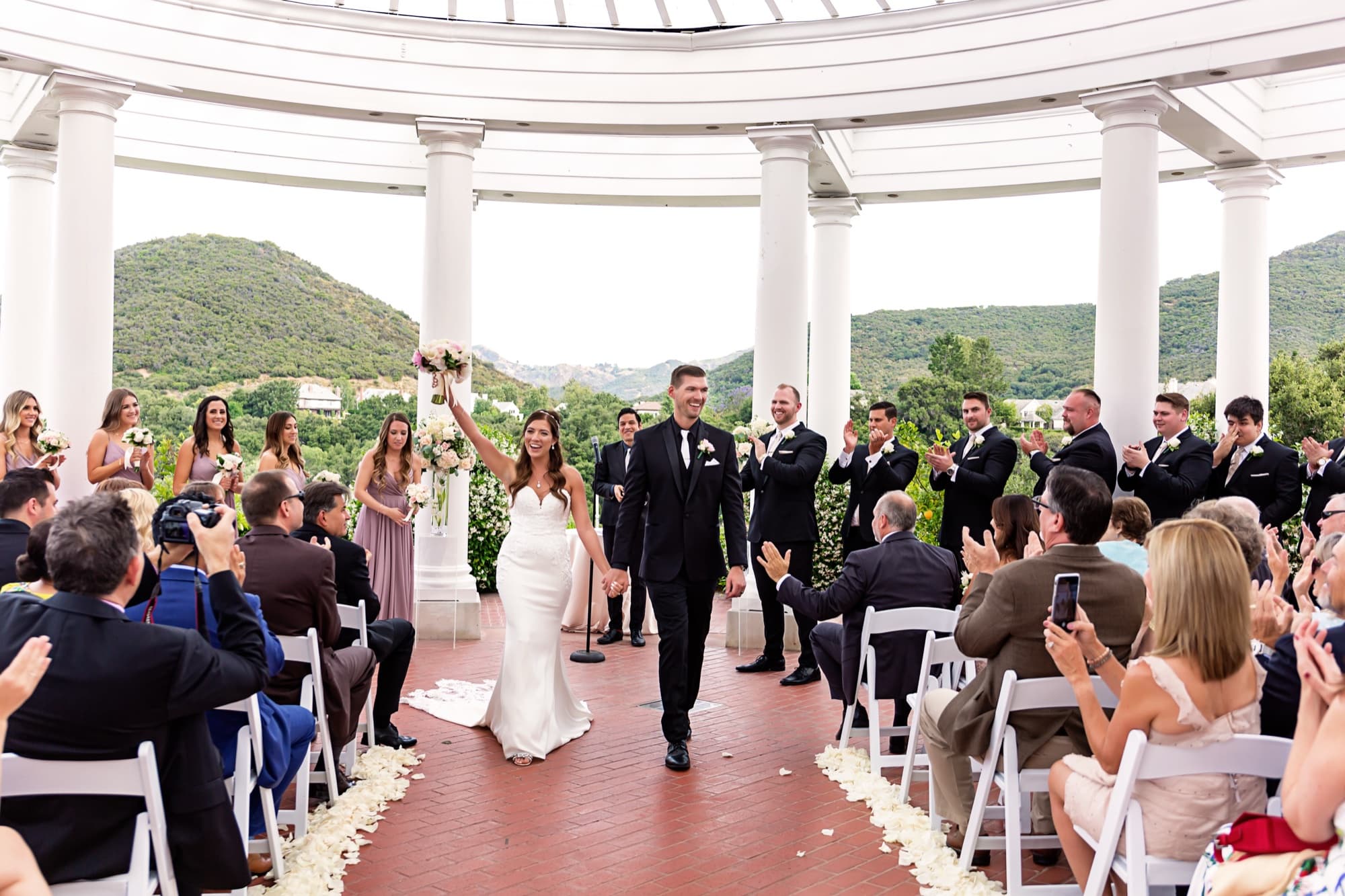 Heather raising her bouquet as she and Brandon walk back up the petal-lined aisle beneath the white rotunda at Sherwood Country Club